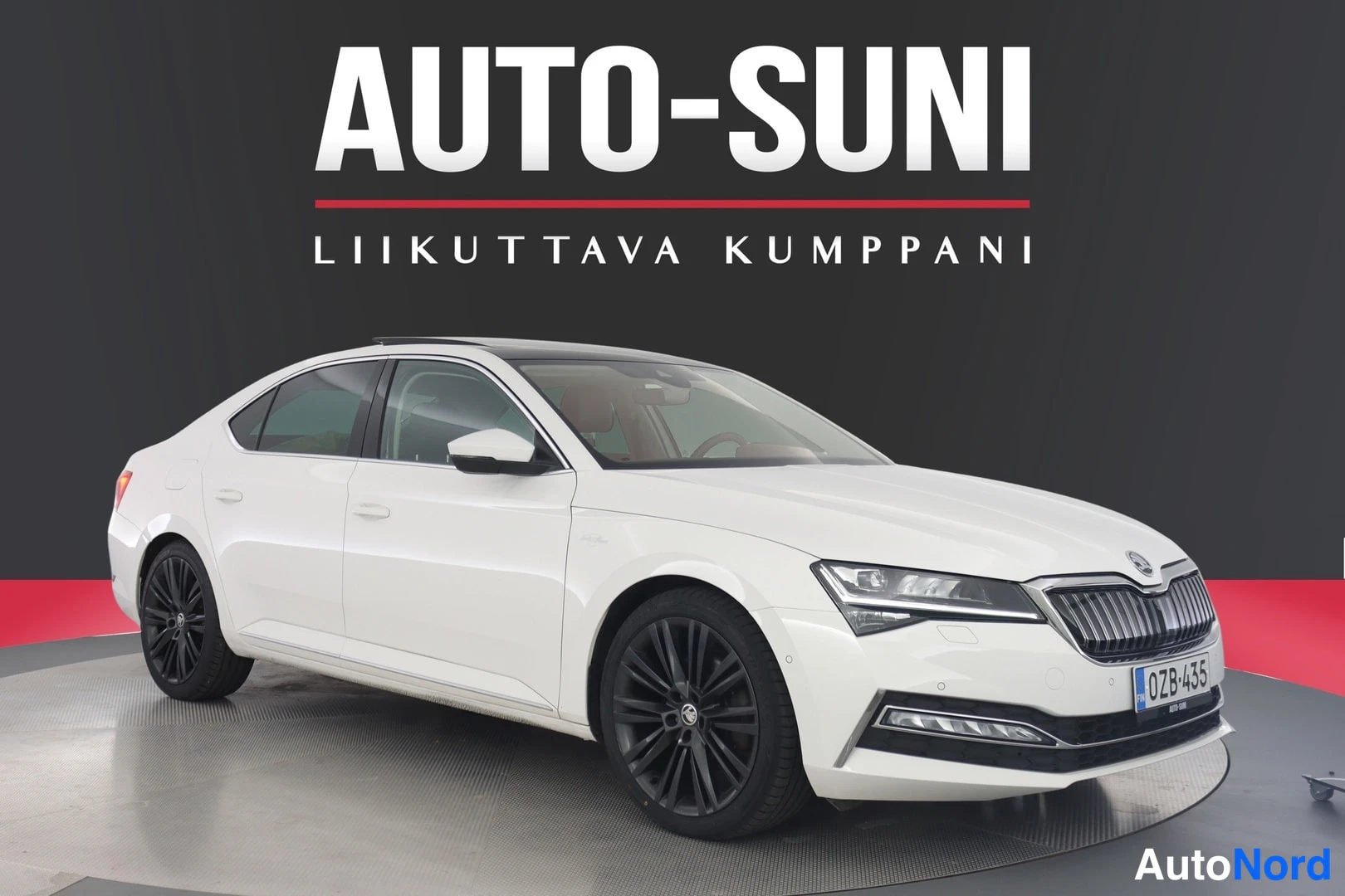 Etuviisto, nokka oikealle: Full side view of a white four-door sedan photographed from the front-left angle in a studio setting with dark gray backdrop and red floor strip; front