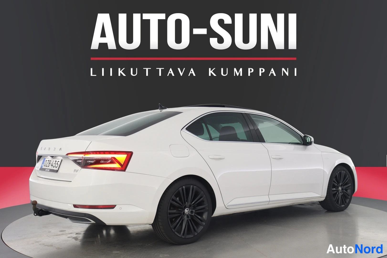 Takaviisto: Auton takakulma valokuvattuna vasemmalta taakse; valkoinen sedan, takavalo syttyneenä, rekisteri, mustat vanteet.