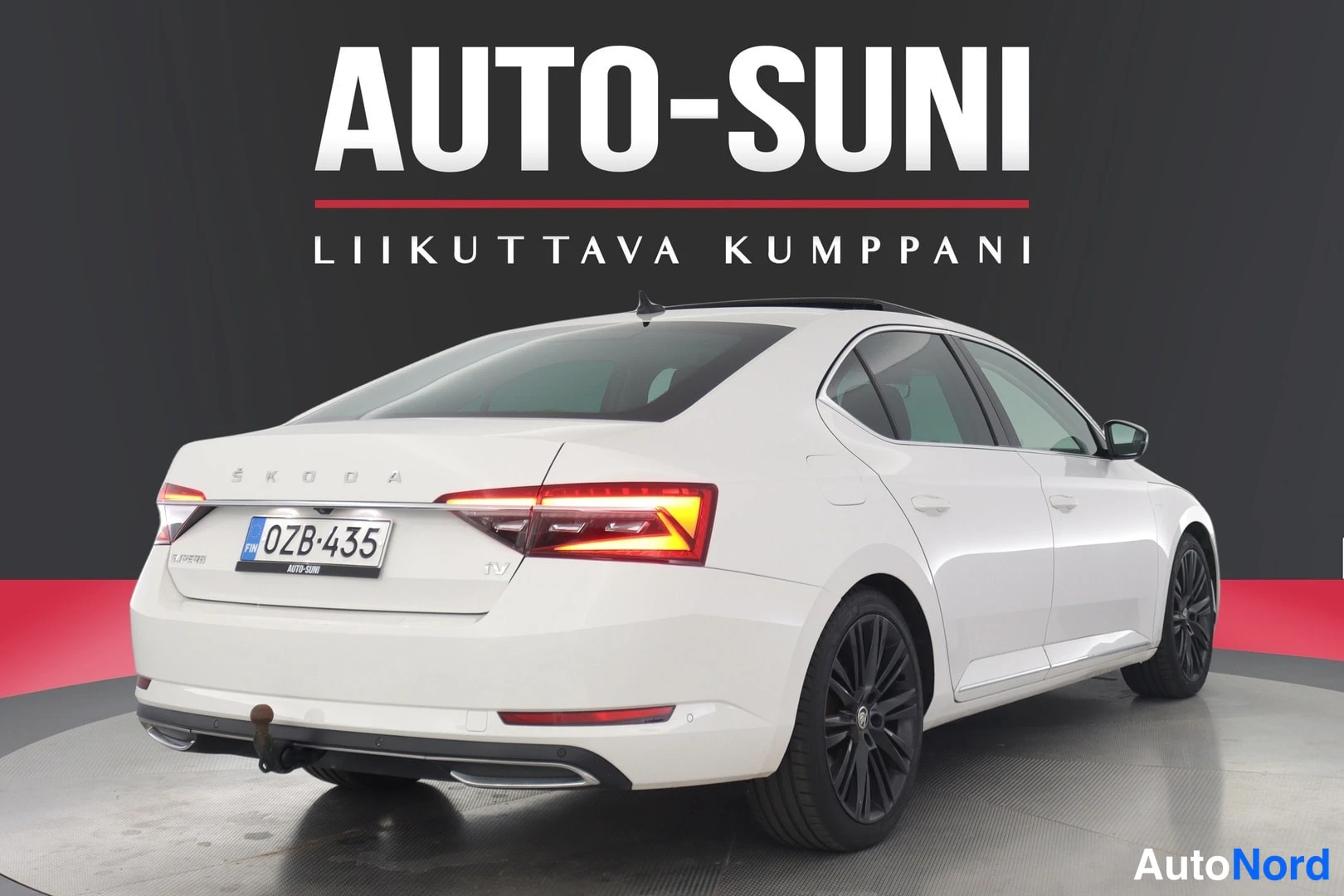 Takaa: Auton takaosa suoraan takaa; valkoinen sedan, ŠKODA-teksti takaluukussa, rekisteri, takavalot syttyneenä.