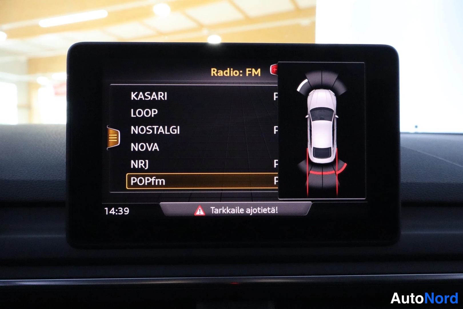 Yksityiskohta: Keskellä näyttö Radio FM -valikolla KASARI, LOOP, NOSTALGI, NOVA, NRJ, POPfm; kartalla ajoneuvon ympäristö; teksti Tarkkaile ajotietä!