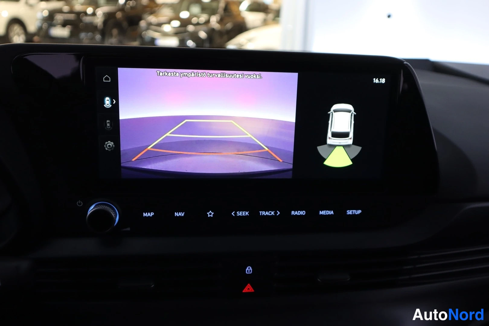Edestä: Kojelaudan infotainment-näyttö näyttää peruutuskameran kuvan pysäköintiviivoin tekstillä 'Tarkasta ympäristö turvallisuutesi vuoksi.' sekä ajoneuvon ympäristönv