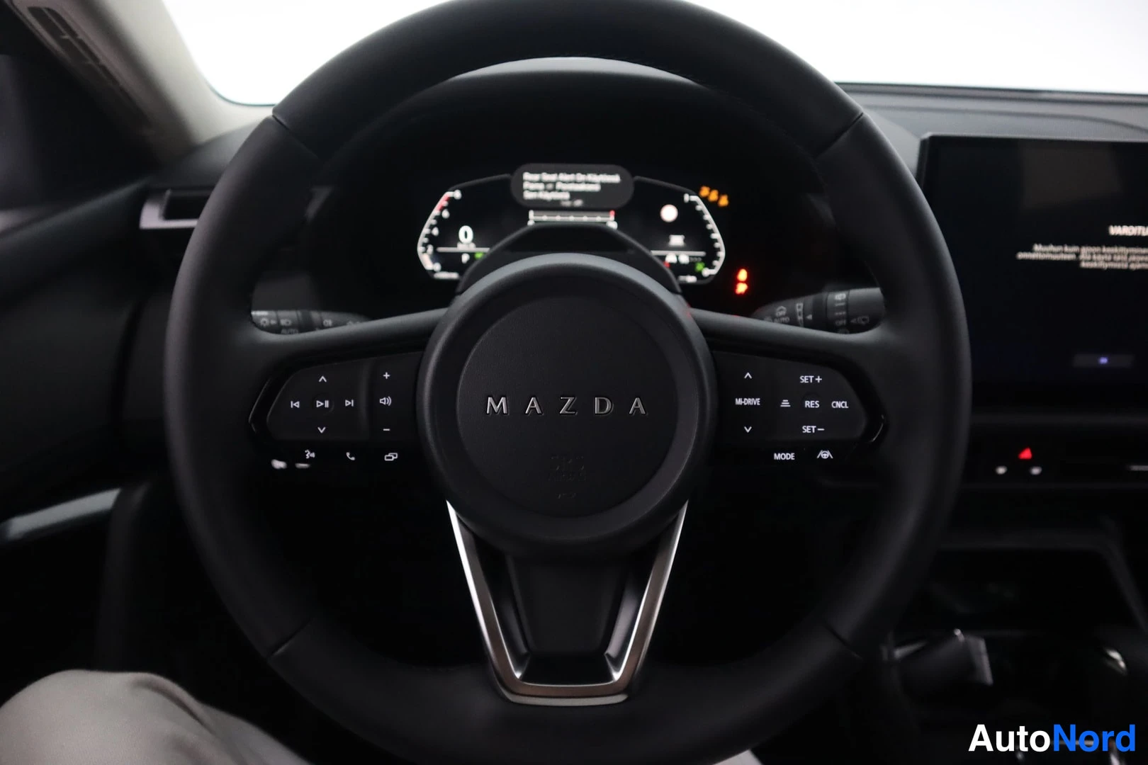 Yksityiskohta: Lähikuva Mazda-ohjauspyörästä, jonka napit ja mittariston näyttö näkyvät; ohjauspyörän napissa MAZDA-teksti ja vasemmalla radio- ja puhelinohjaimet.