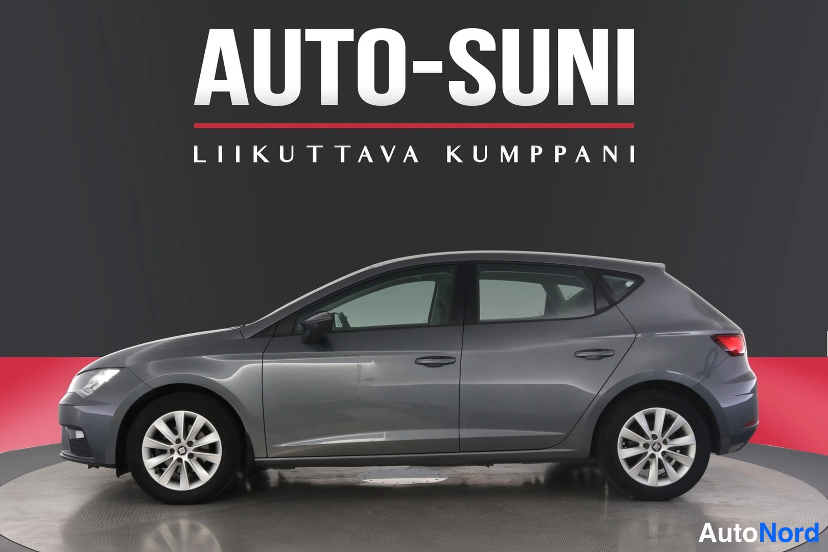 Sivulta: Harmaa viistosti sivusta kuvattu Seat Leon -henkilöauto studio----ympäristössä, Auto-Suni-mainos taustalla.