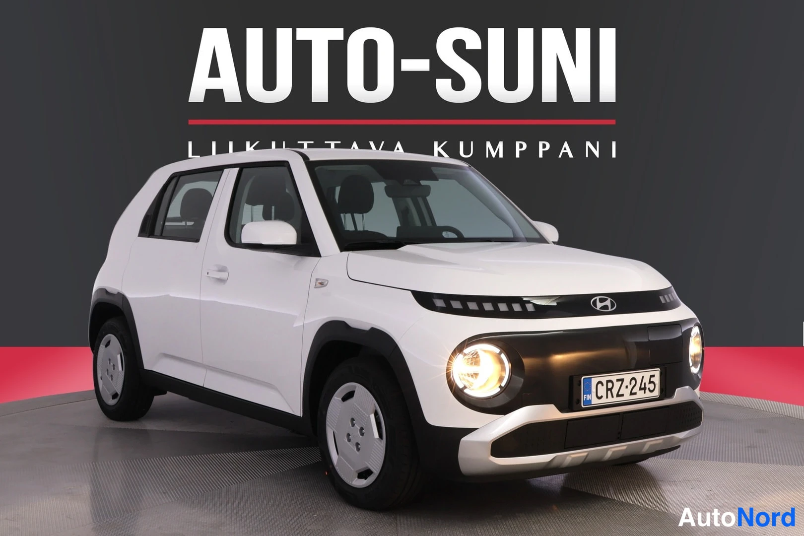 Etuviisto, nokka oikealle: Valkoinen Hyundai Inster -henkilöauto etukulmasta oikeaan etuosaan kuvattuna studio­ympäristössä, etuvalot päällä.