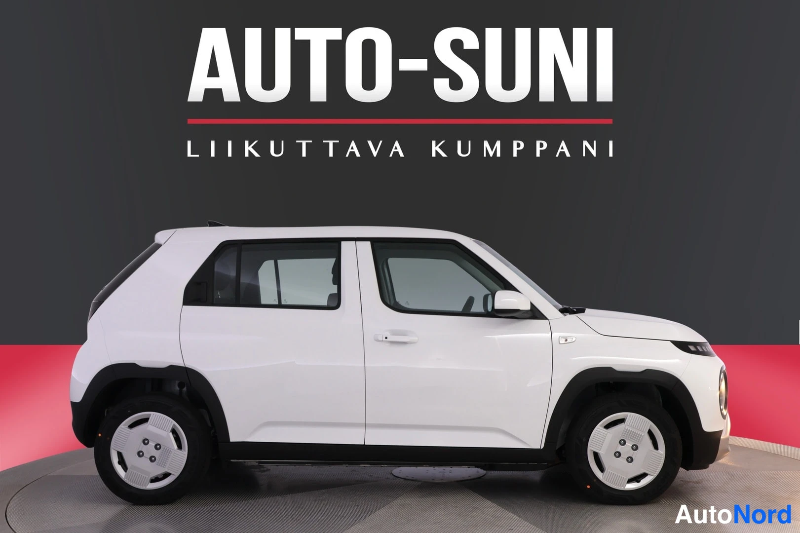 Etuviisto, nokka oikealle: Valkoinen pieni viisiovinen auto sivuprofiilikuvassa harmaalla-punaisella studiotaustalla; yläosassa Auto-Suni-logo ja teksti 'Liikuttava kumppani'. Etuv