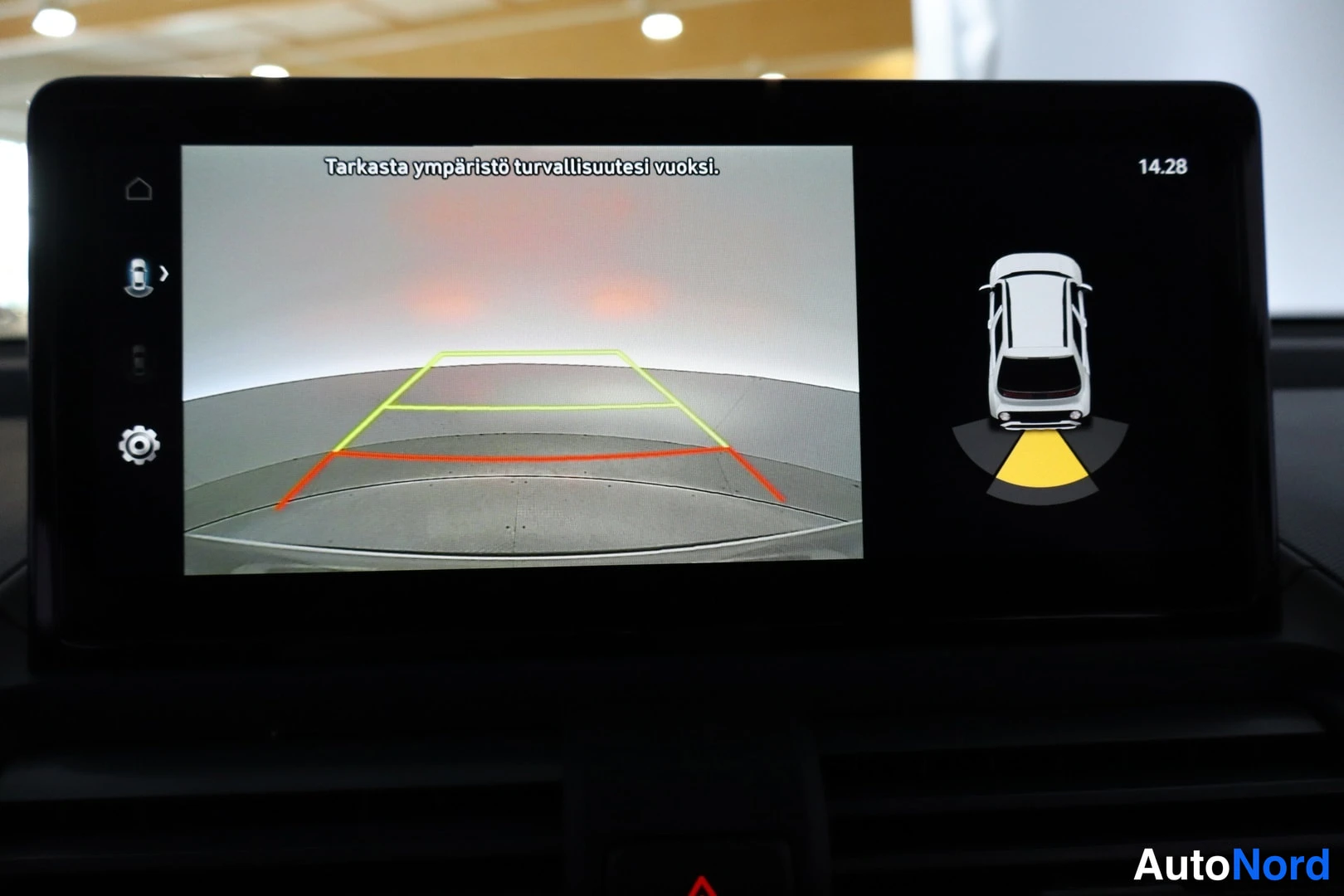 Muu: Infotainment-näyttö näyttää peruutuskamerakuvan ohjausviivoilla ja tekstillä 'Tarkasta ympäristö turvallisuutesi vuoksi.'