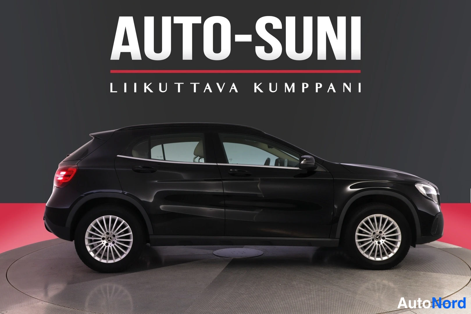 Sivulta: Kuvassa on musta Mercedes-Benz GLA -kattoauto valkoisella sisätiloilla. Auto on sijoitettuna kiertopyörälle, ja kuvan oikealla puolella näkyy Auto-Suni -merkki.