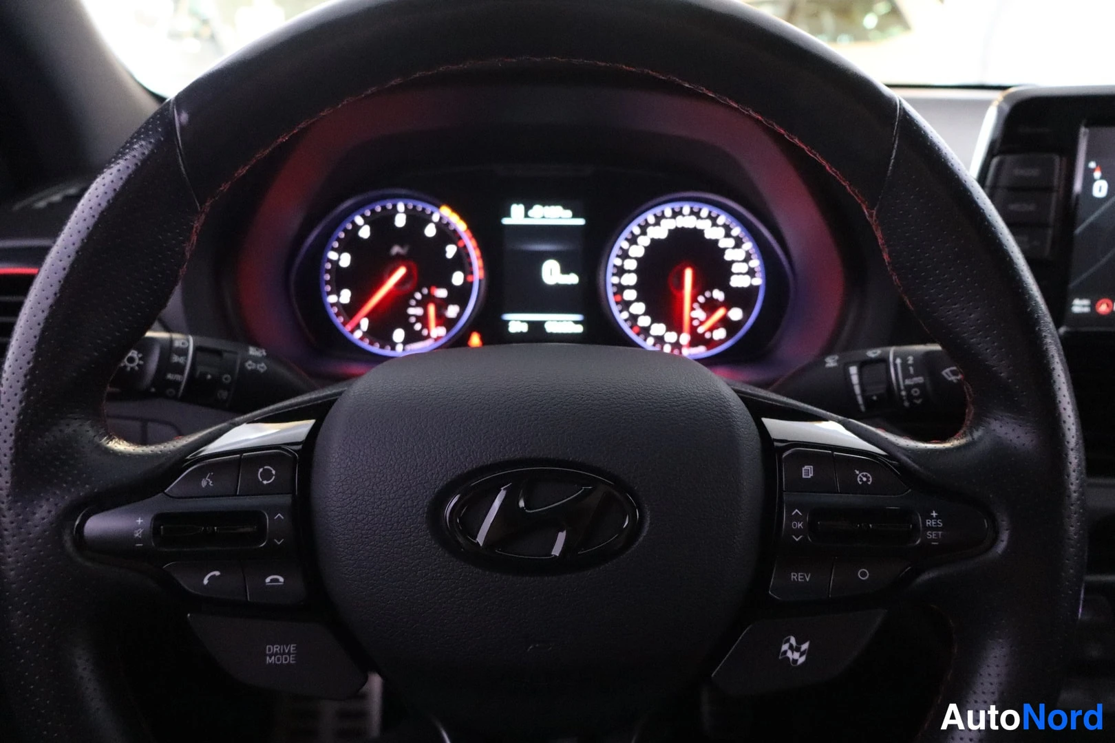 Sivulta: Sisäkuva ohjauspyörästä ja kojelaudasta: Hyundai-logo napissa, tumma nahkainen vanne punaisin ompeluin, analoginen tahdonomittari ja nopeusmittari, digitaalinäy