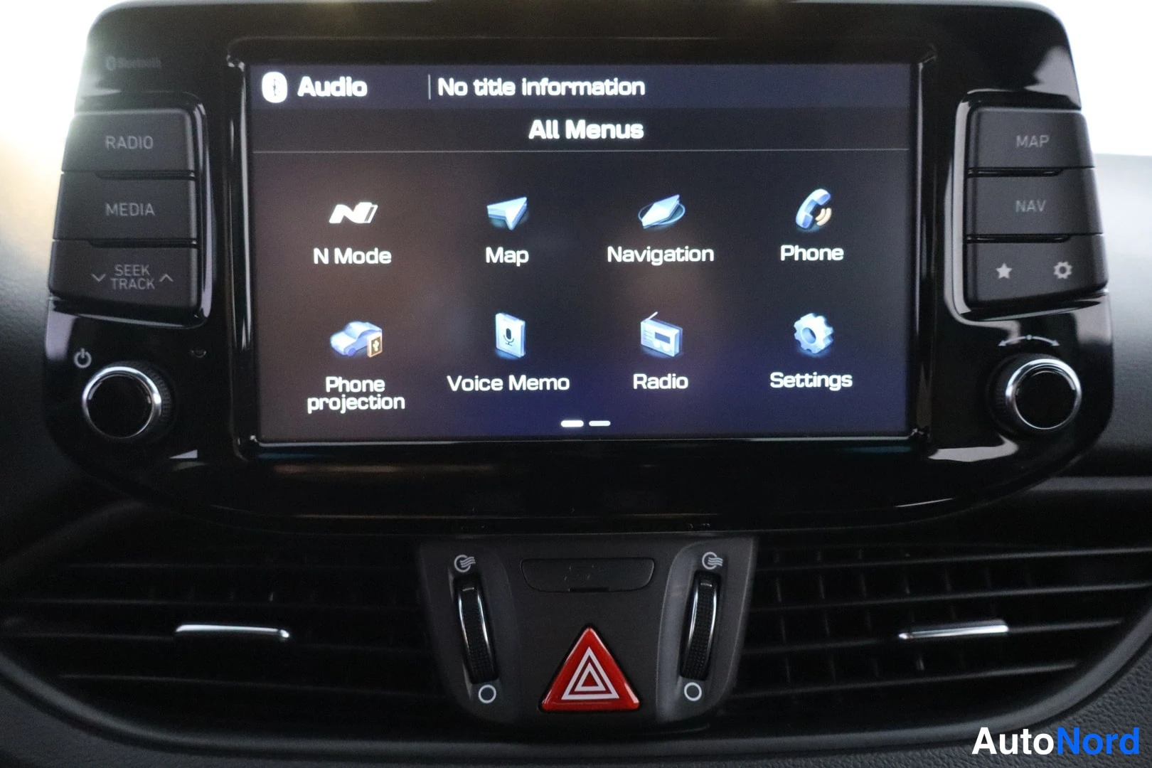 Etuviisto, nokka vasemmalle: Auton keskusnäytön valikko: Audio, N Mode, Map, Navigation, Phone, Phone projection, Voice Memo, Radio ja Settings; näytön alla punainen varoituskolmio