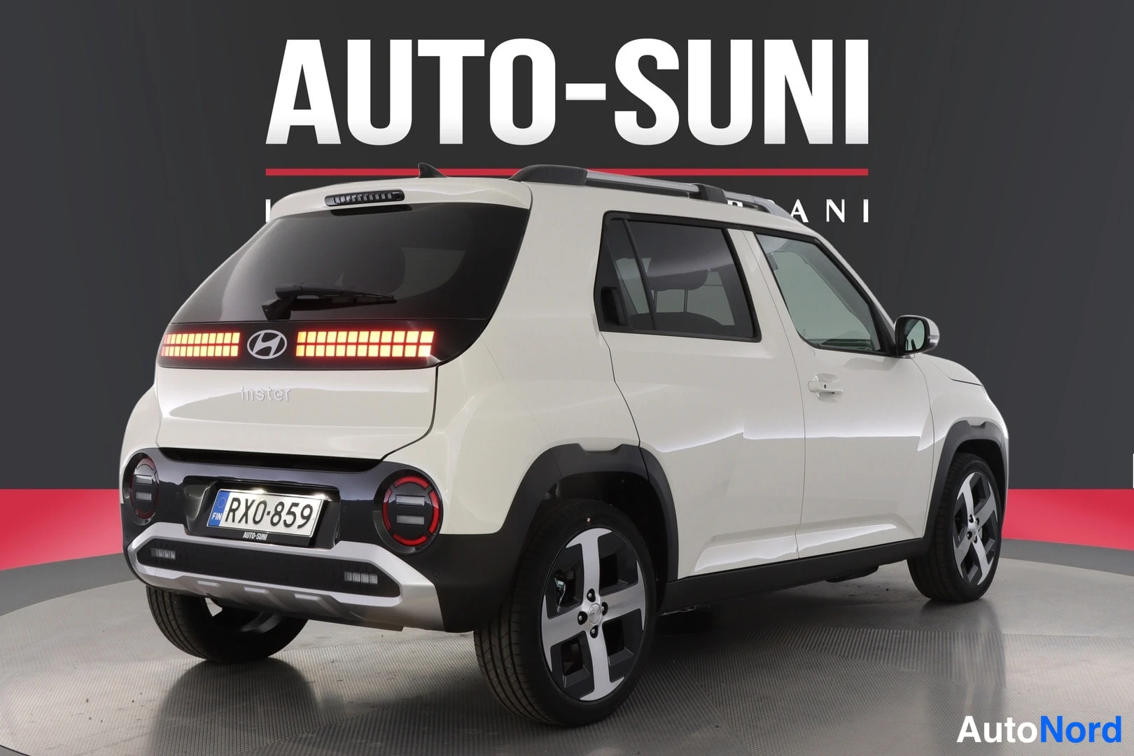 Takaviisto: Rear three-quarter view of the white car from the right side. The rear tail lights are illuminated in red, along with the central brake light bar. Roof rails an