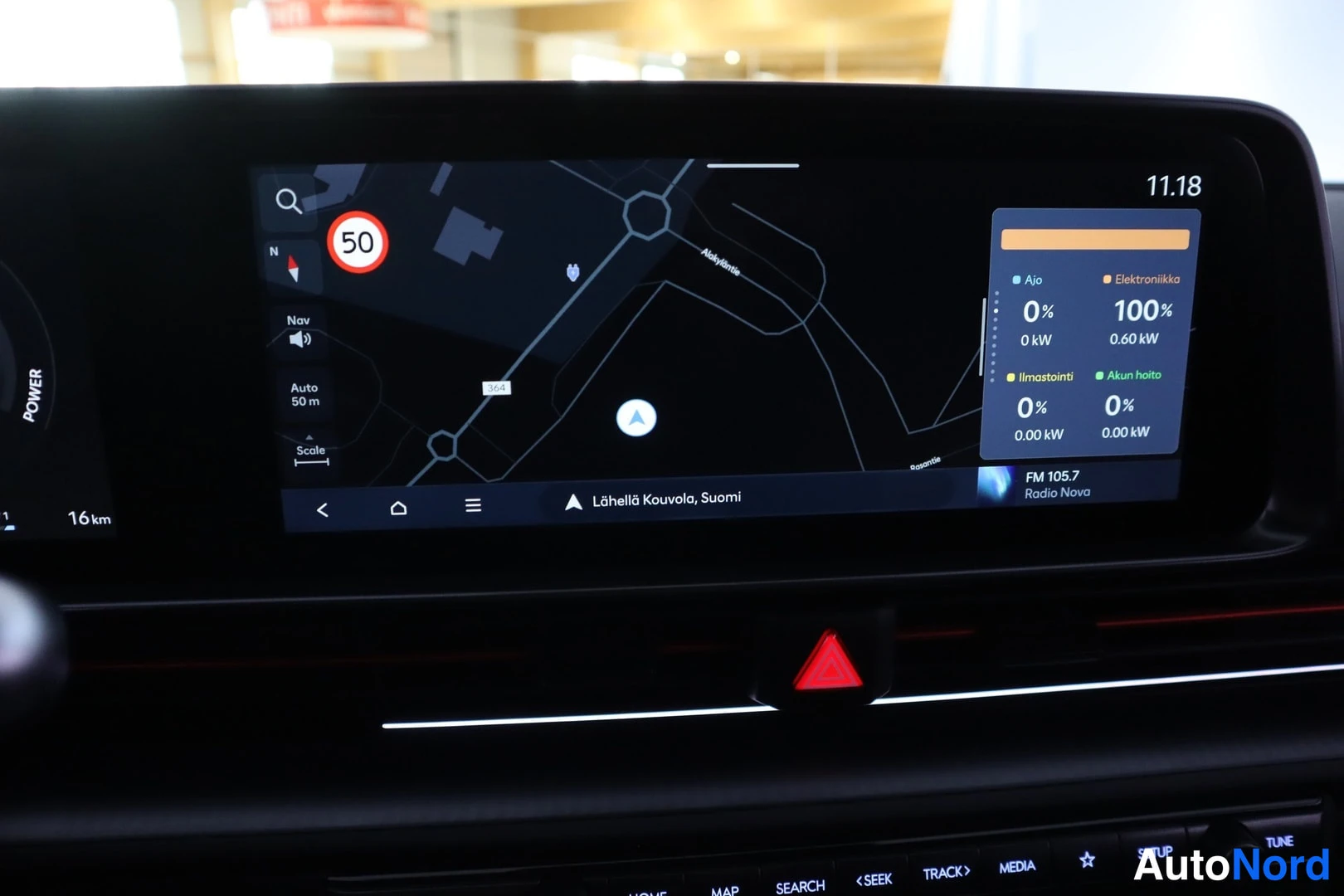 Edestä: Infotainment-näytön lähikuva: navigointikartta tekstillä 'Lähellä Kouvola, Suomi', 50-nopeusrajoitusmerkki ja lataustilapaneeli (Ajo 0 %, Elektroniikka 100 %, I