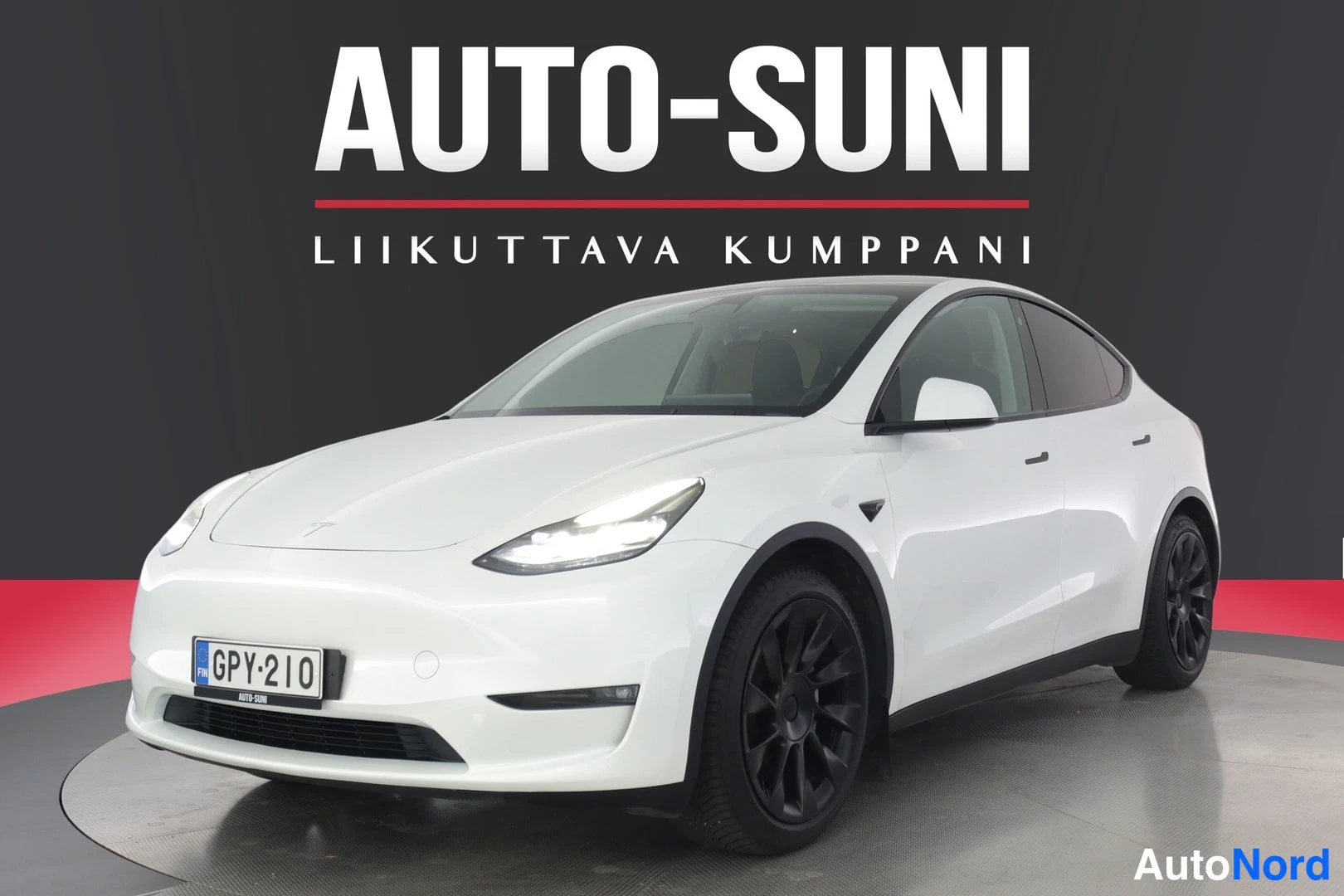 Etuviisto, nokka vasemmalle: Valkoinen Tesla Model Y etuviistosta, keula kuvan vasemmalla puolella; ajovalot päällä, mustat vanteet, rekisterikilpi.