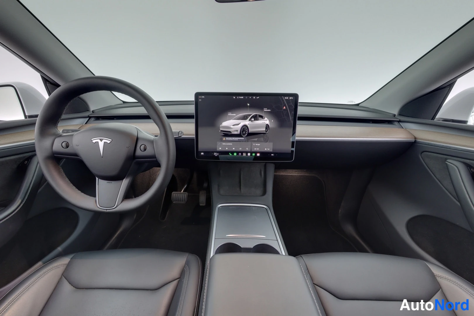 Muu: Tesla Model Y:n musta ohjaamo: Tesla-merkillinen ohjauspyörä, iso keskellä sijaitseva kosketusnäyttö, puunväriset koristelistat ja mustat penkit.
