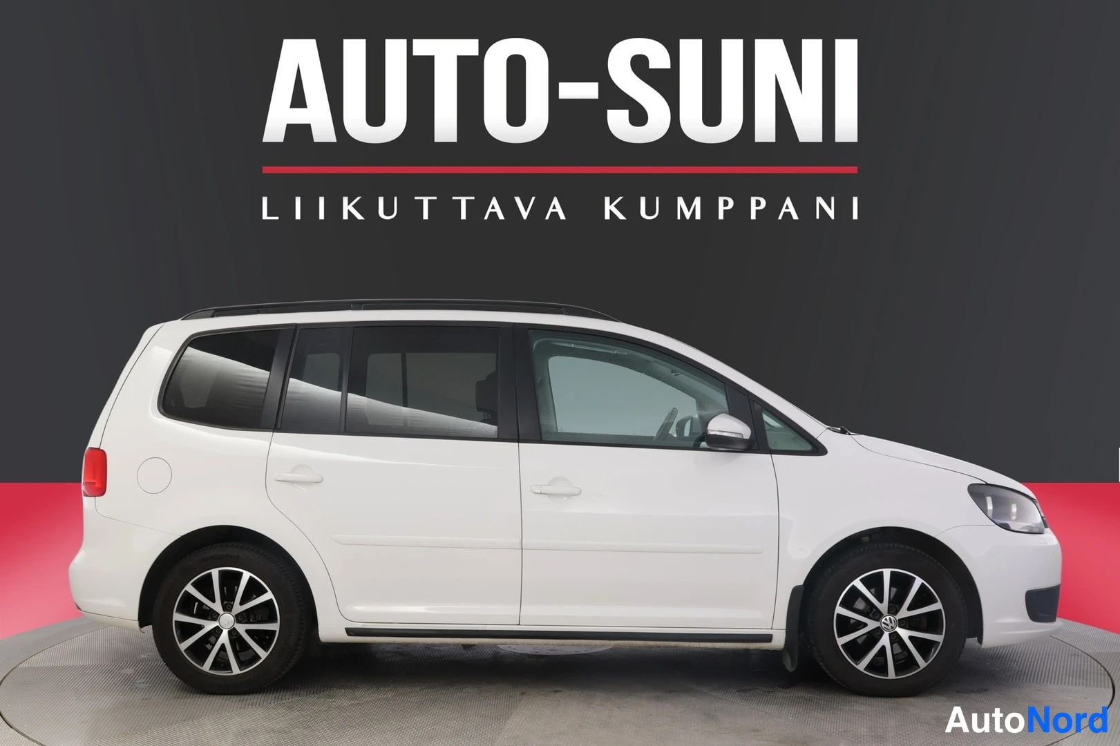 Sivulta: Oikean puolen sivunäkökulma valkoisesta Volkswagen-autosta. Huomioitavaa on auton katossa olevat kattohoidot ja renkaat. Taustalla on tumma seinä ja lattialla p