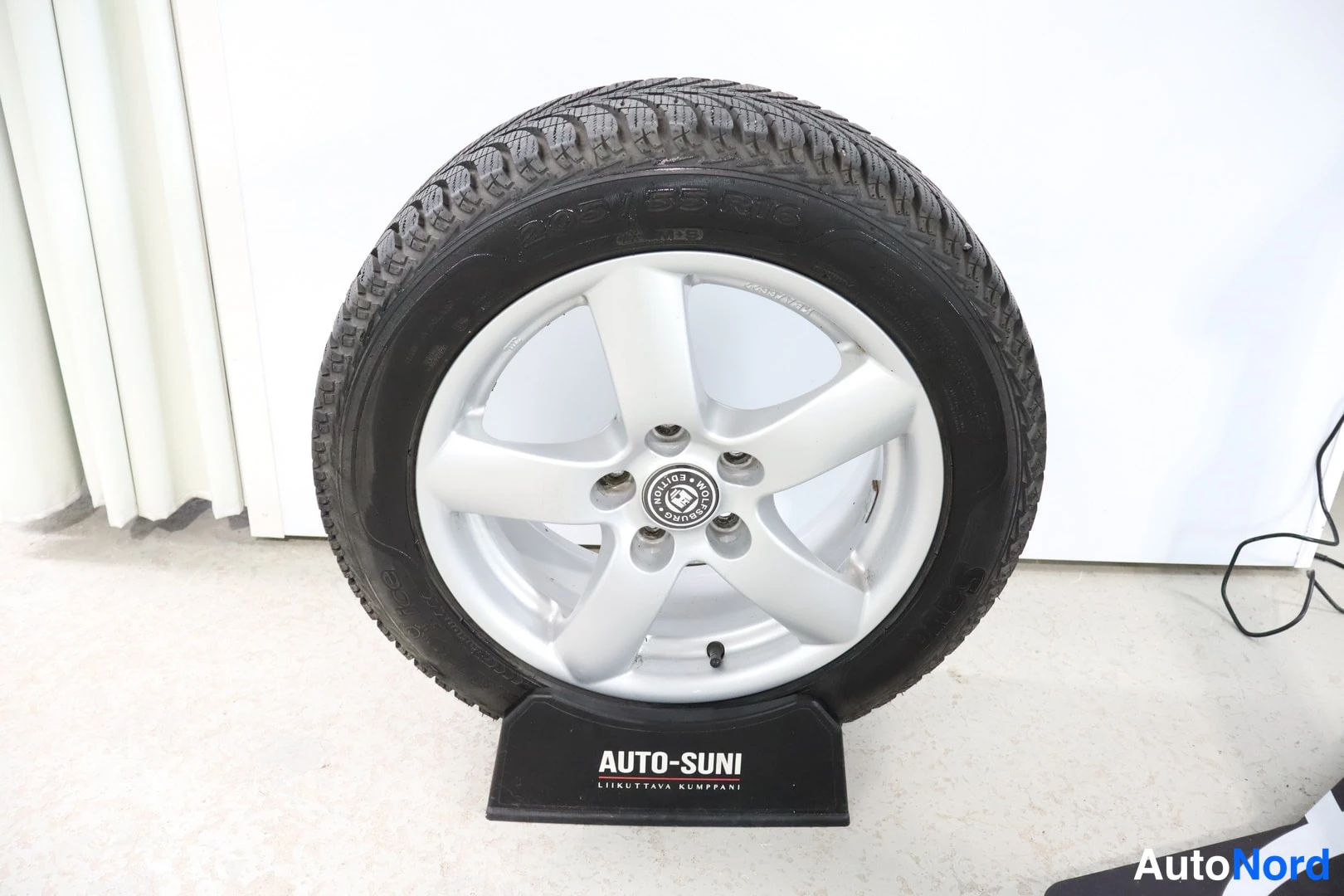 Yksityiskohta: A silver five-hole aluminium alloy wheel with a winter tyre mounted on it, displayed upright on a black stand reading AUTO-SUNI.