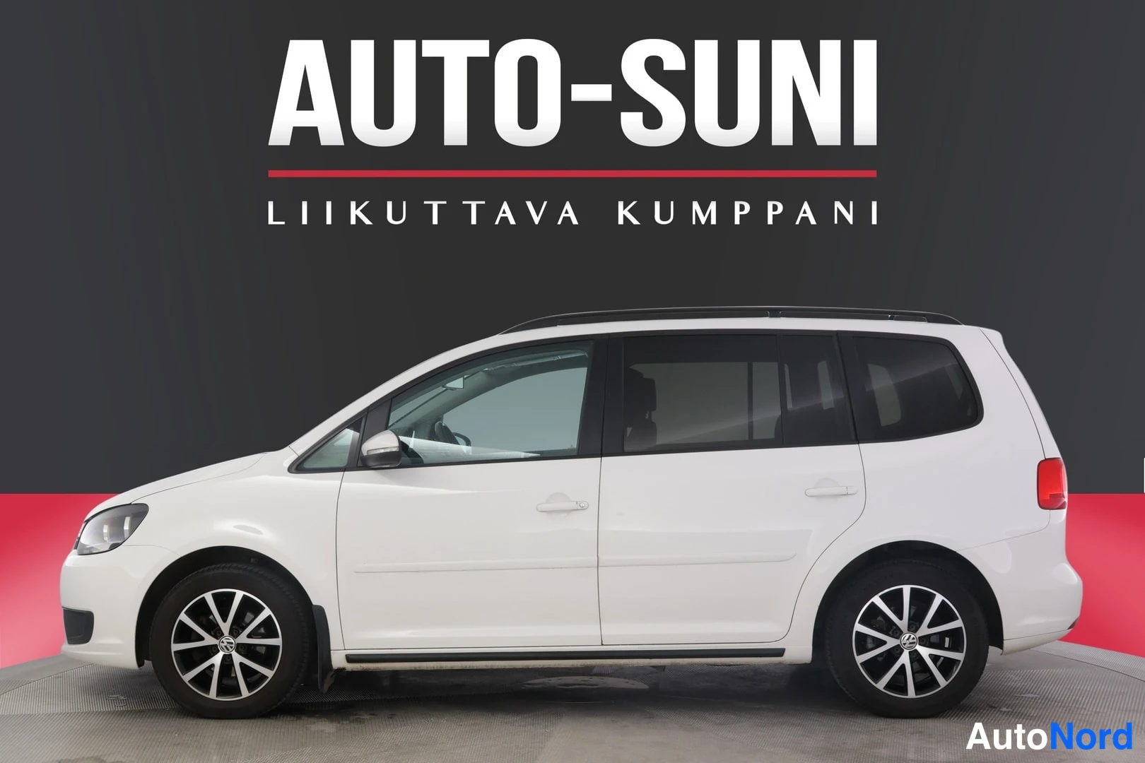 Sivulta: Valkoinen Volkswagen Touran seisoo varastossa vasemmalta sivulta kuvattuna. Autossa on kattoharjat ja renkaissa volkkarin logot.