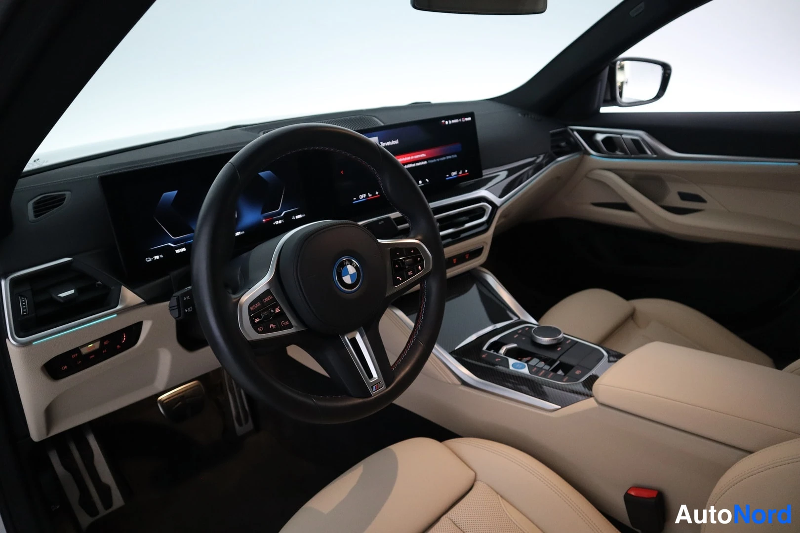 Edestä: Henkilöauton ohjaamo: musta M-merkillinen urheiluohjauspyörä BMW-emblemillä, kaareva kaksoisnäyttö kojelautaan, beige nahkaverhoillut penkit ja keskikonsolin iD