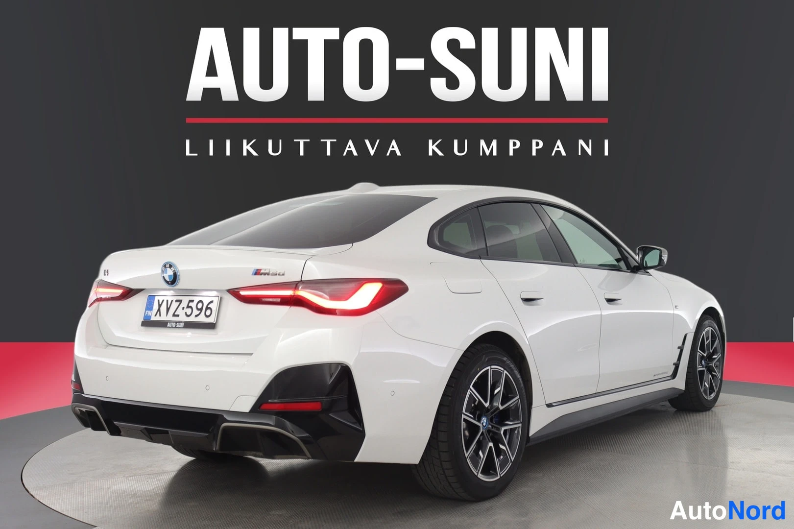 Takaviisto: Valkoinen BMW i4 lähes suoraan takaa vasemmasta viistosta; takaluukussa BMW-merkki ja M50-tunnus, perävasen takavalo syttyneenä, rekisterikilpi.