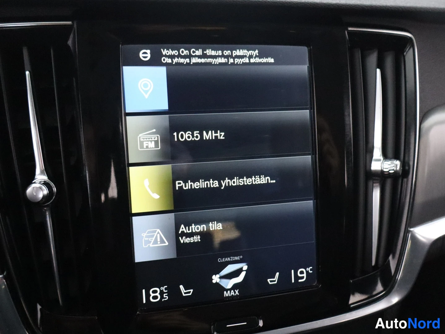 Ohjaamo: Close-up of the vertical central infotainment screen between two vertical air vents. Screen displays a menu with icons and text including radio frequency 106.5