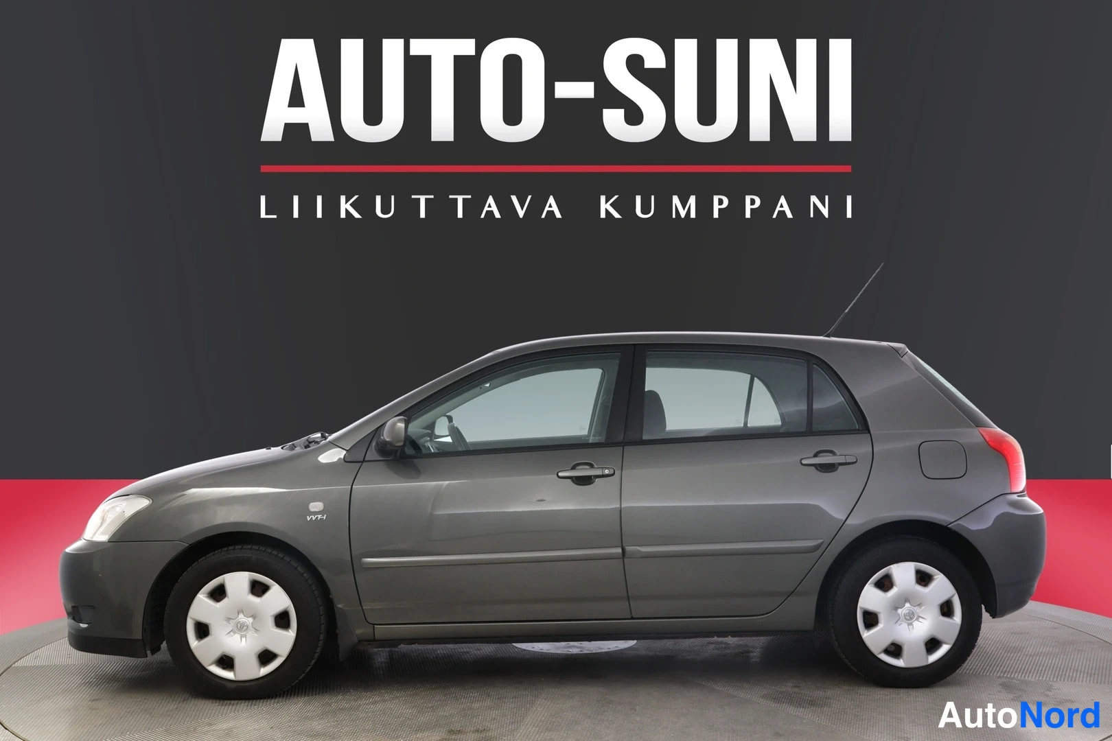Etuviisto, nokka vasemmalle: Autosuni-kauppiaan logolla taustalla tummanharmaa Toyota Corolla -henkilöauto kuvattu etukolmesta, keulan osoittaen vasemmalle.