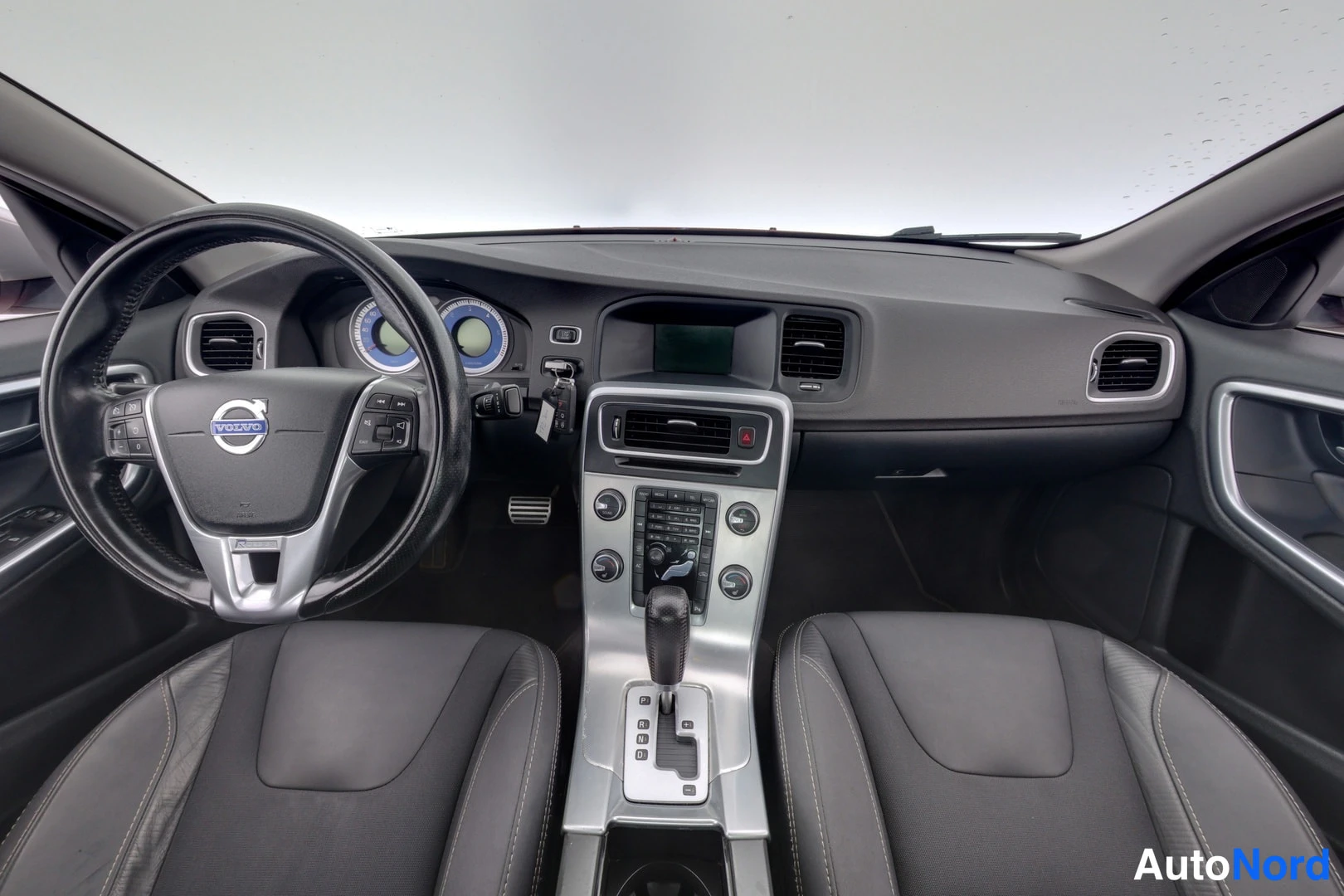 Muu: Volvo S60:n ohjaamo etusat alta kuvattuna: musta mittaristo sinisillä taustoilla, Volvo-merkitty ohjauspyörä, keskikonsoli ja kaksi tummaa etuistuinta.