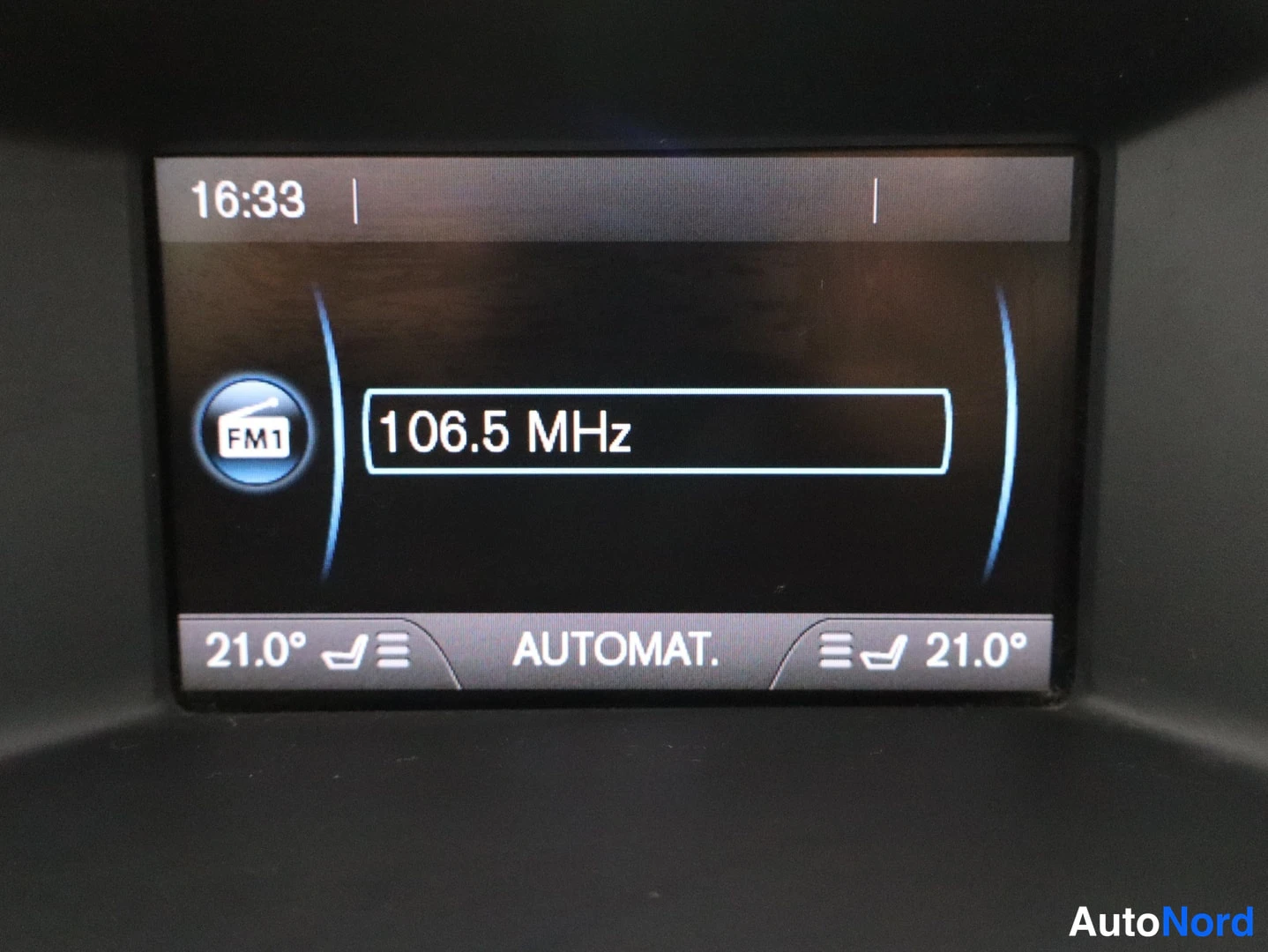 Yksityiskohta: Auton infonäyttö näyttää kelloa 16:33, radioasemaa FM1 106.5 MHz ja molempien lämpötilan 21,0 astetta, tekstillä AUTOMAT.