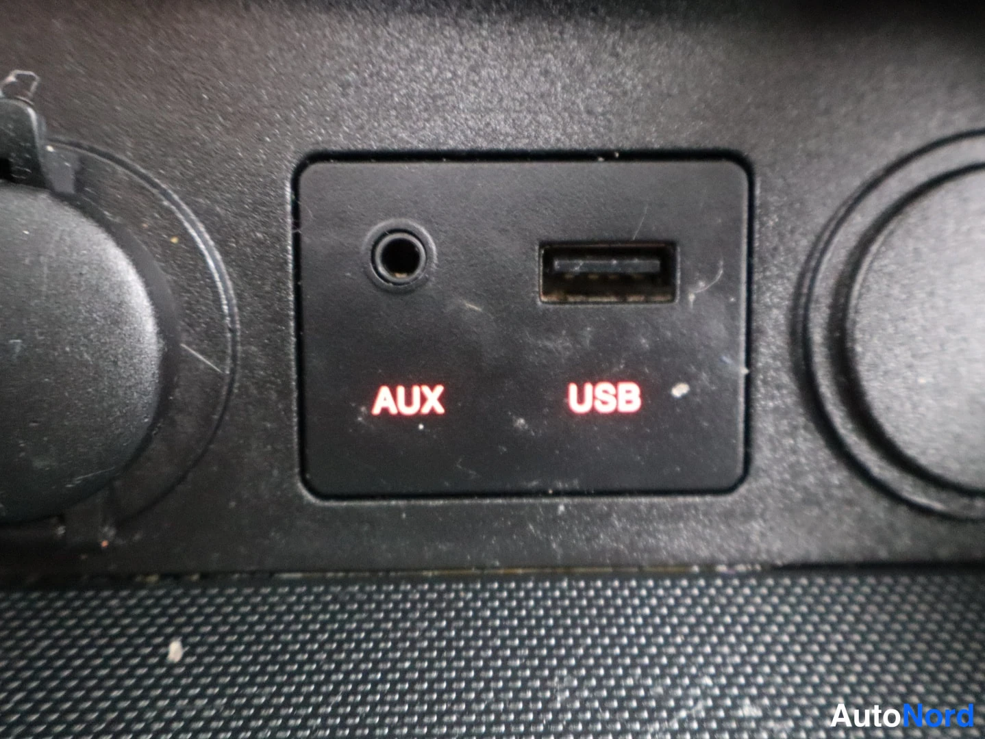 Muu: Sisätilakuva: AUX- ja USB-liitännät mustassa paneelissa, punaisella valaistut tekstit AUX ja USB, molemmin puolin pyöreät aukot.