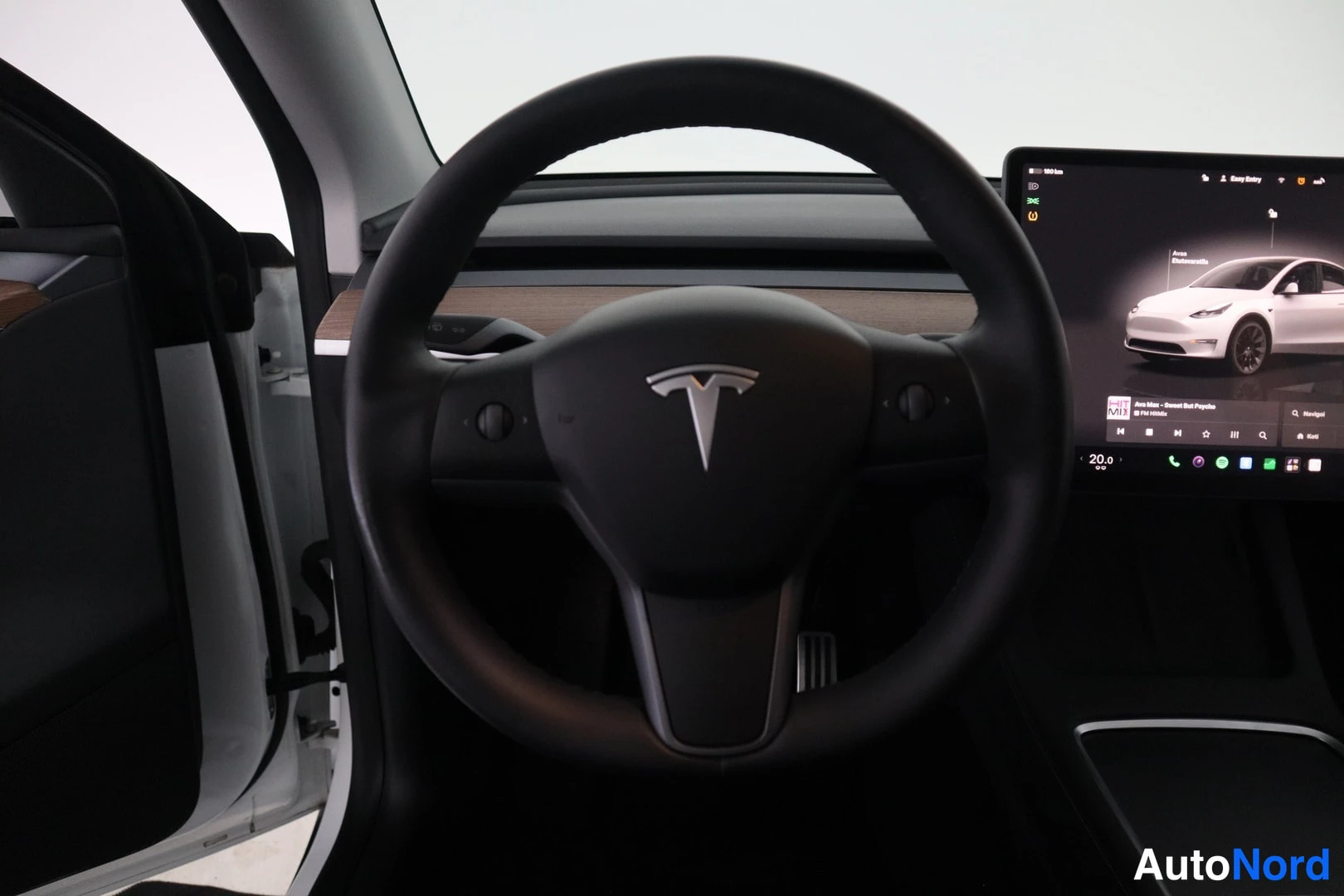 Yksityiskohta: Ohjauspyörän läheiskuva Tesla-logoa ja kojelaudan keskinäyttöä vasten. Näytössä näkyy auton kuva ja valikko.