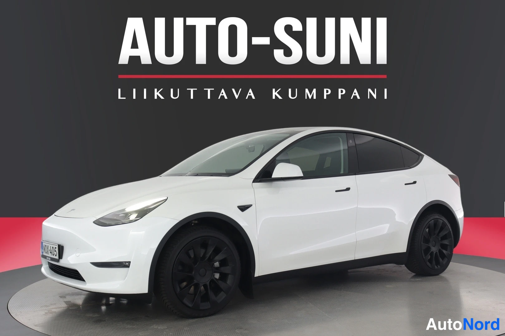 Etuviisto, nokka vasemmalle: Valkoinen Tesla Model Y -henkilöauto kuvattu kolmivartalokulmassa auton nokka vasemmalla, mustat vanteet, harmaa tausta.