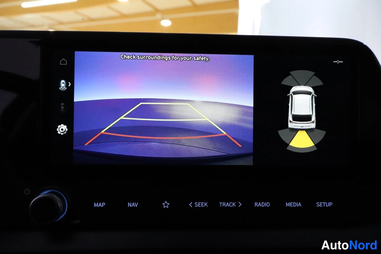 Yksityiskohta: Infotainment-näyttö näyttää peruutuskameran kuvan ohjausviivoineen ja tekstin 'Check surroundings for your safety.'