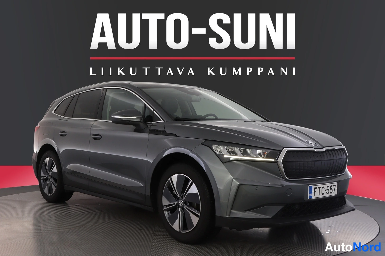 Etuviisto, nokka oikealle: A metallic grey vehicle photographed from a front three-quarter angle on a rotating studio display against a dark wall with 'AUTO-SUNI' signage.