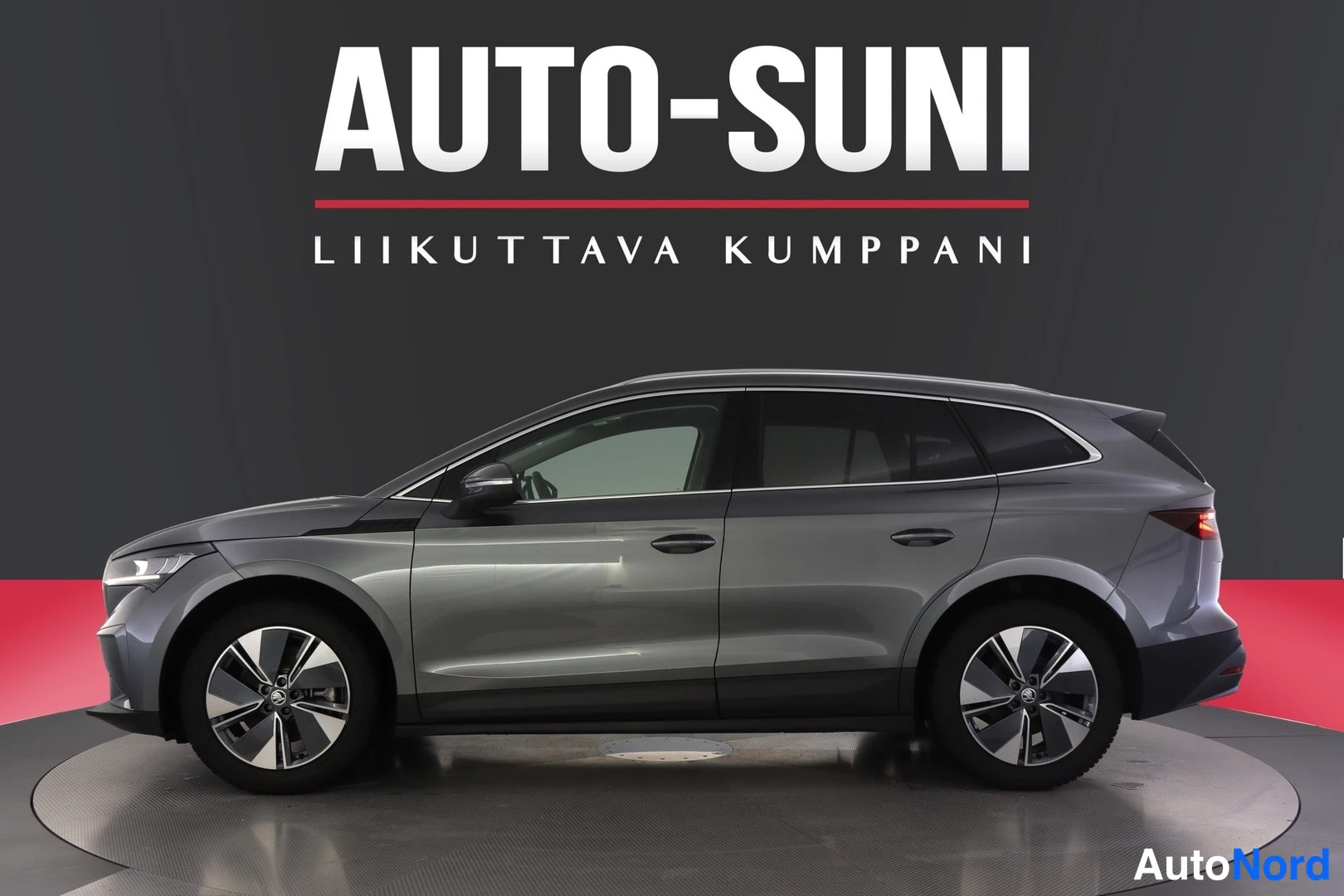 Sivulta: Ajoneuvon sivuprofiili: tummanharmaa farmarimallinen auto rekisterikilpi vasemmalla, myyjän logot tekstinä taustalla.