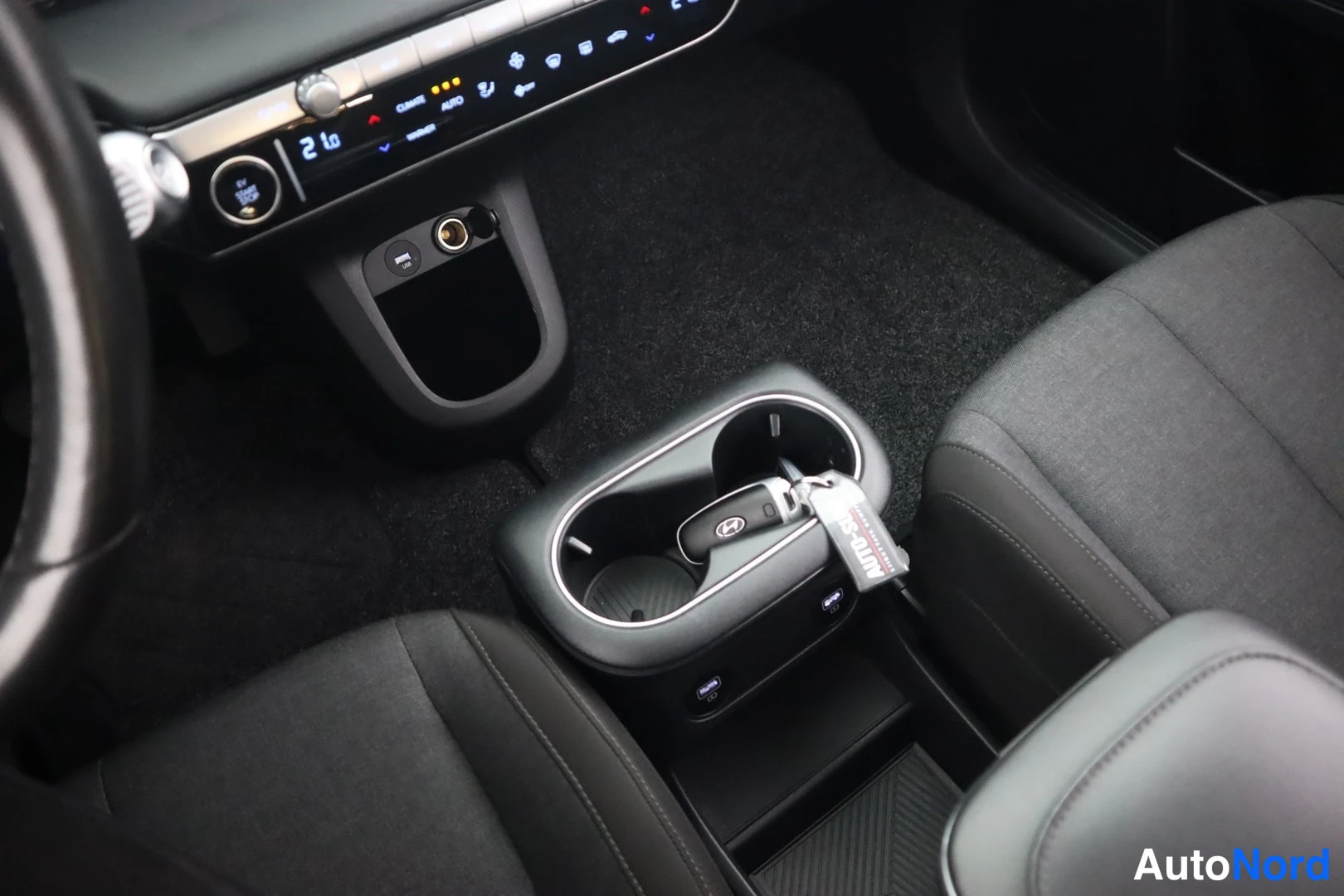 Yksityiskohta: Näkymä auton sisälle: keskikonsolin kupitelineessä Hyundai-avain AUTOPRO-rannekkeessa, taustalla ilmastointipaneeli ja tummat kudosistuimet.