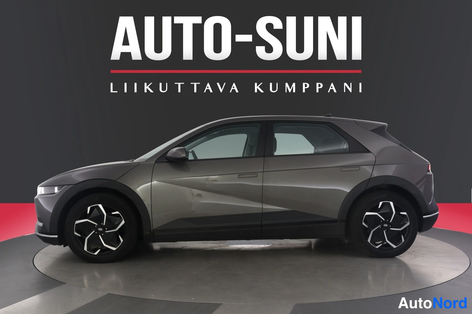 Sivulta: Hyundai IONIQ 5 -henkilöauton etuvasemmosta kolmasosaa näyttävä kuva autoliikkeen studiossa, autossa harmaa maalipinta ja tummat alumiinivanteet.