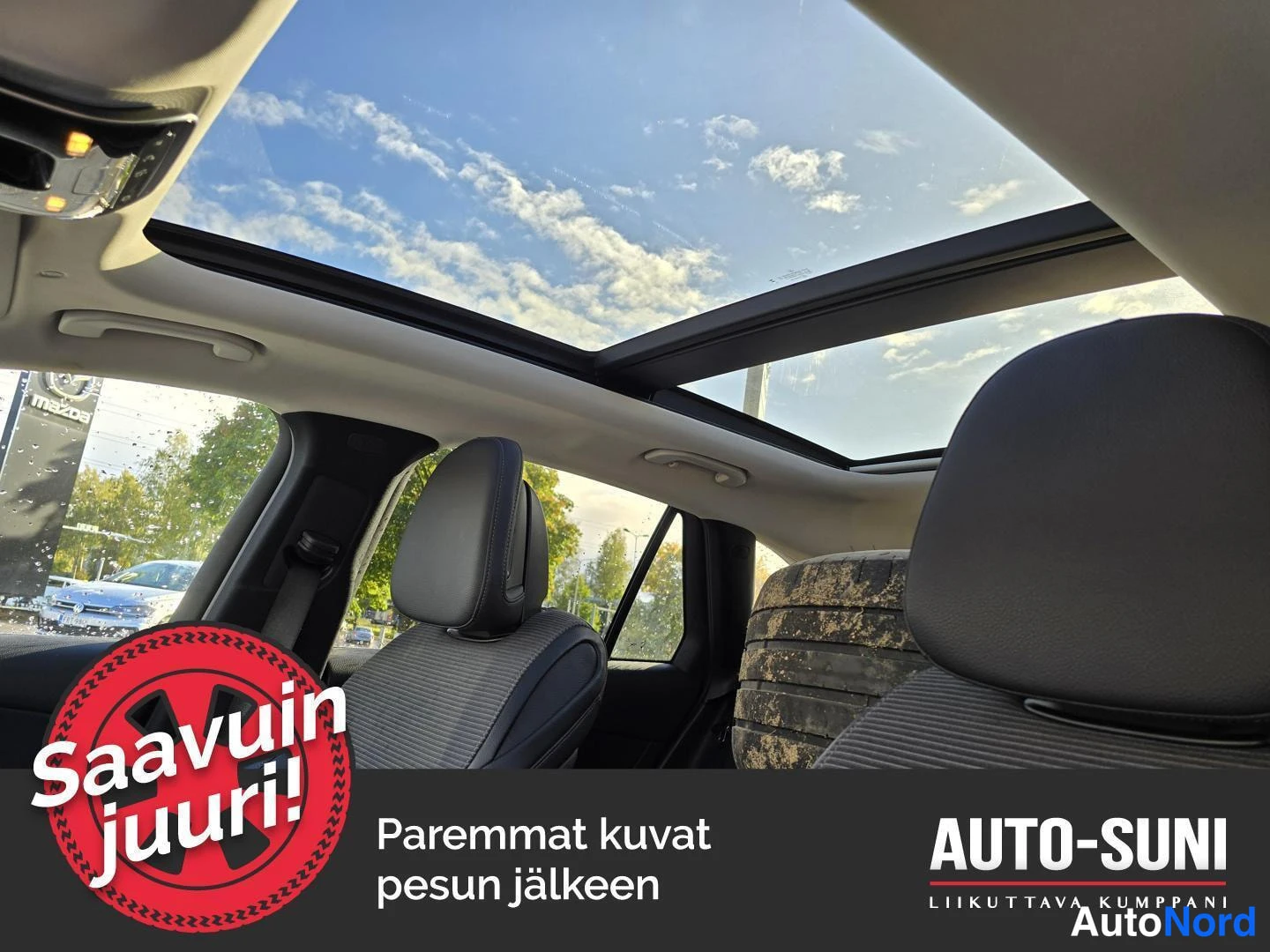 Etuviisto: Auton sisätilat: avattava panoraamakatto, jonka läpi näkyy sininen pilvinen taivas, sekä etupenkkien pääntuet ja musta kattokonsoli.