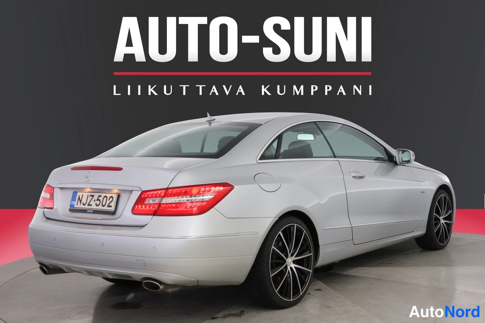 Takaviisto: Mercedes-Benz-coupén takakolmeneljänneskuva, hopeanvalkoinen koritys, takavalot ja rekisterikilpi, mustat vanteet, kuvan yläosassa AUTO-SUNI-logo.