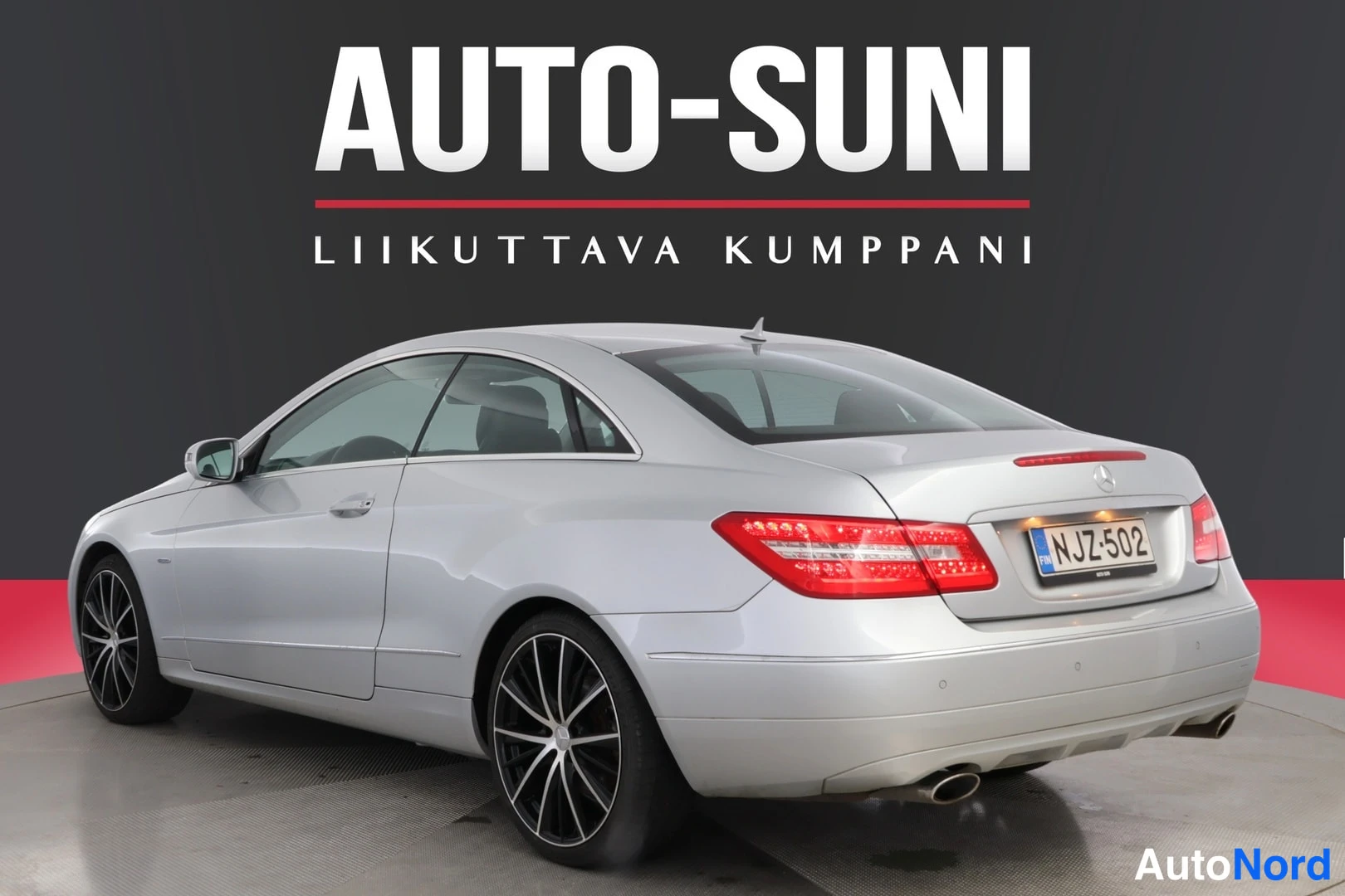Takaviisto: Mercedes-Benz-coupén takakolmeneljänneskuva vasemmalta takaa, hopeanvalkoinen koritys, takavalot ja rekisterikilpi, mustat vanteet, kuvan yläosassa AUTO-SUNI-lo