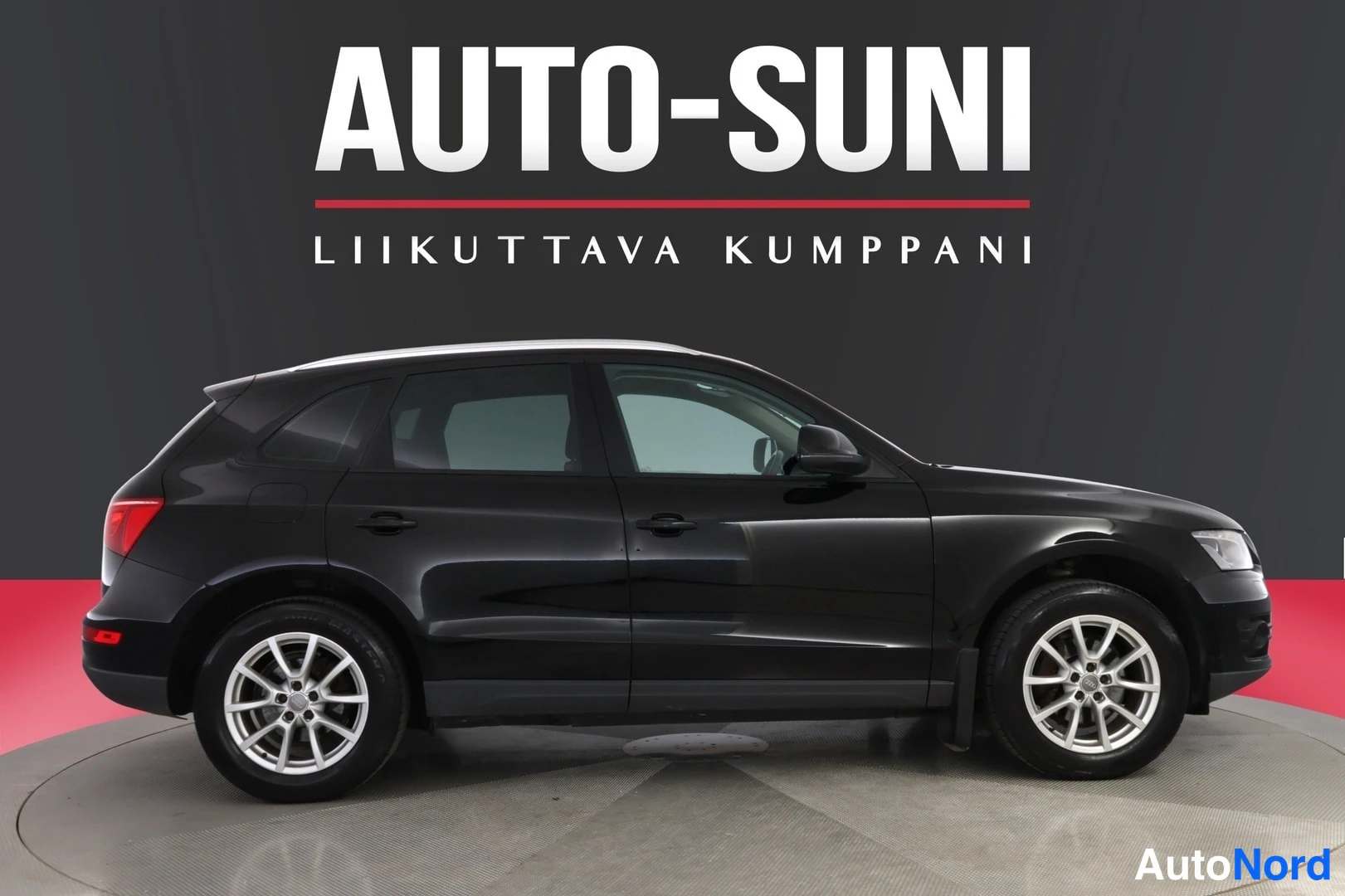 Etuviisto, nokka oikealle: Mustan Audi Q5 -maasturin etukulmanäkyvä sivukuva studio-oloissa, ympyräplatformilla, taustalla liikkeen logo.