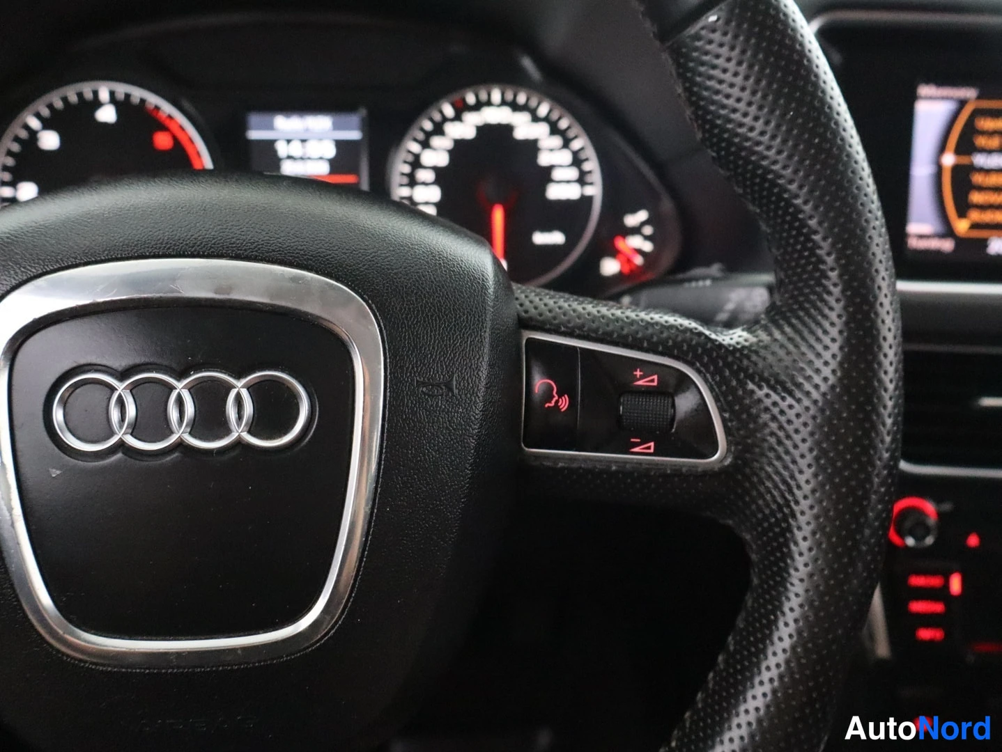 Yksityiskohta: Audi-ohjauspyörän oikea kytkinpainike puhelin- ja äänenvoimakkuusmerkeillä, taustalla mittaristo.