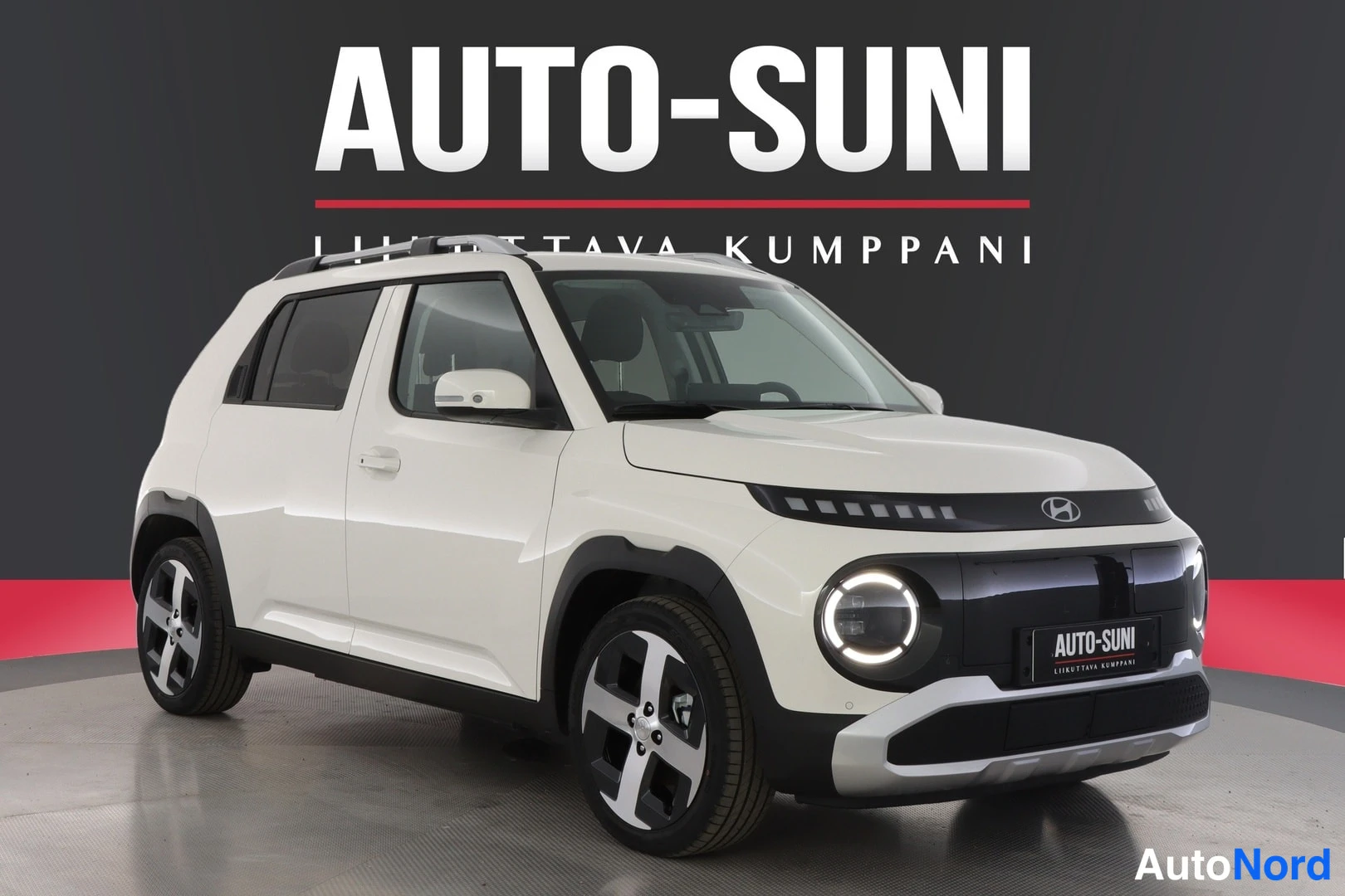 Etuviisto, nokka oikealle: Valkoinen Hyundai Inster pieni sähkö-SUV esillä studion pyörivällä alustalla, kolmenNeljännesnäkymä etupuolelta ja oikealta sivulta; näkyvissä kevytmetal
