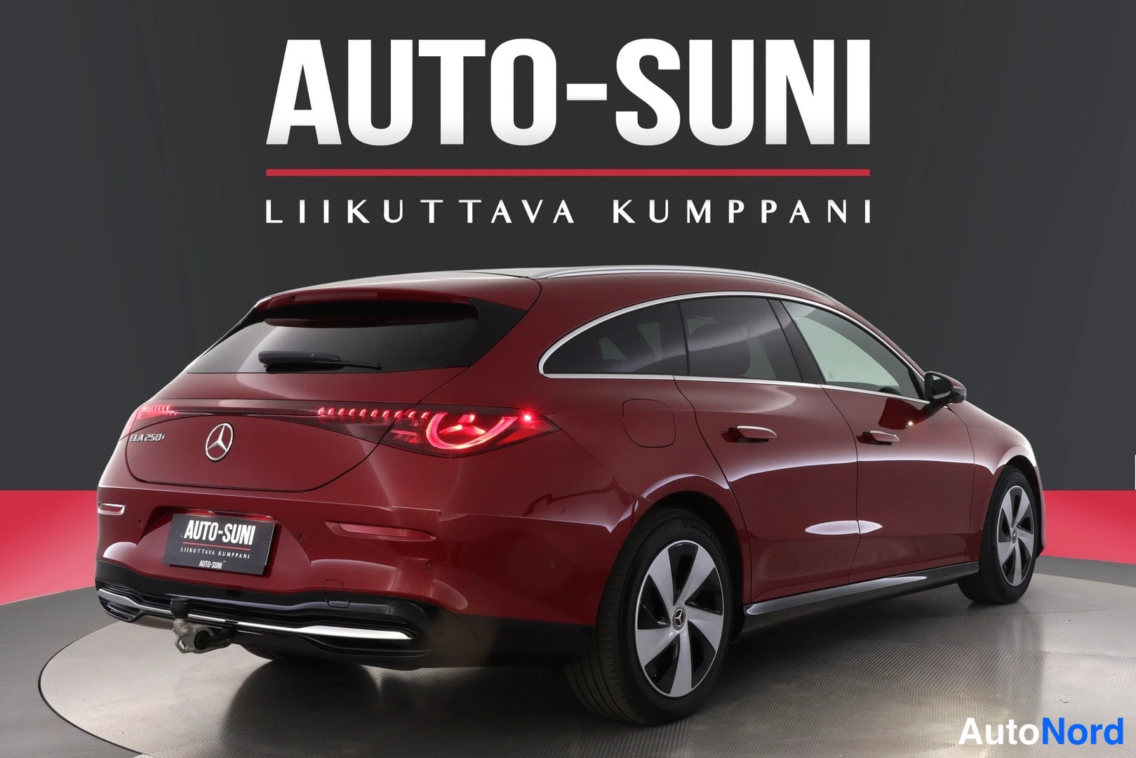 Takaviisto: Tummanpunainen Mercedes-Benz CLA -farmari takaaviistosta, perä kuvan vasemmassa laidassa, takavalot palavat, studiotausta.