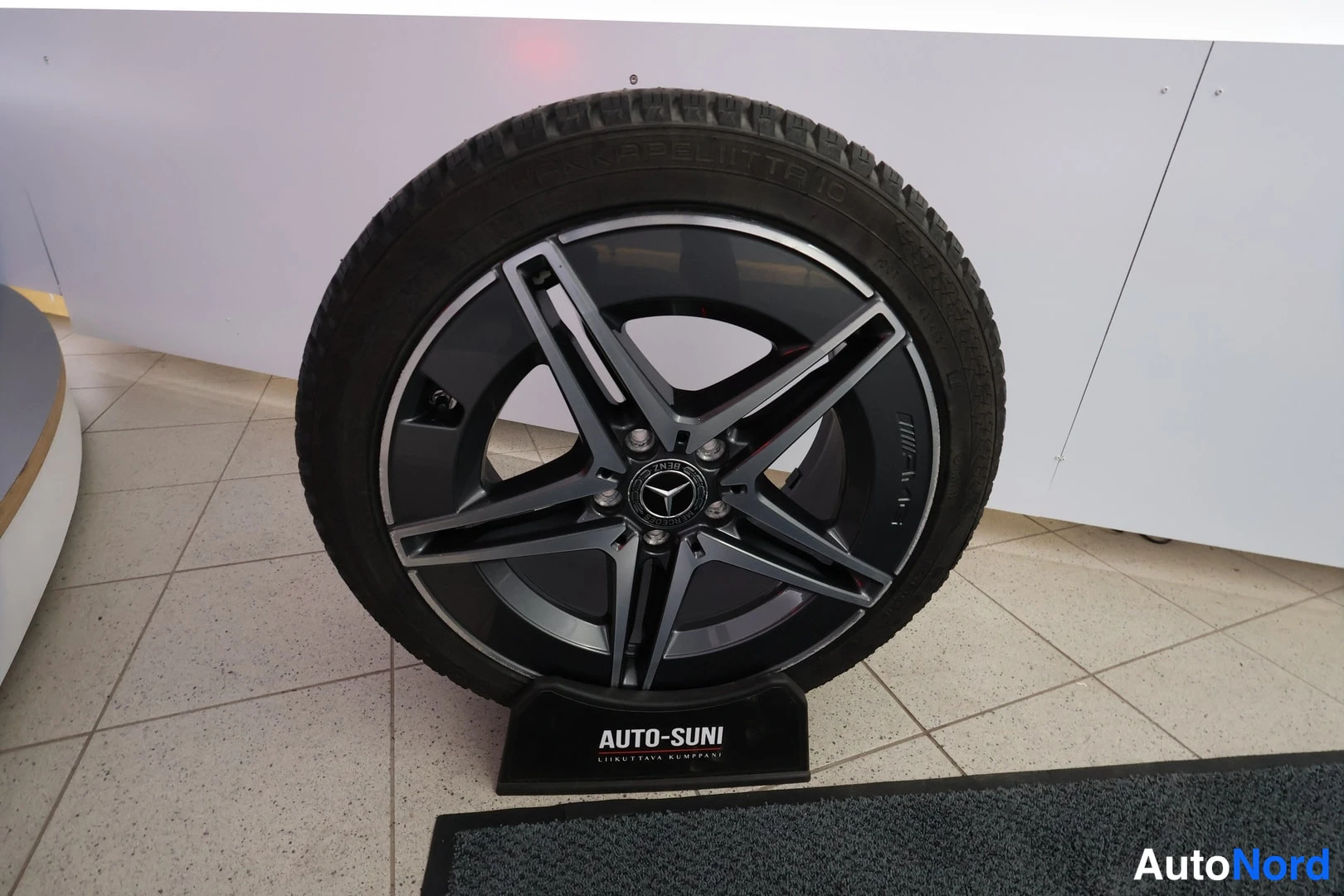 Yksityiskohta: Sivuvarapyörä näkyvissä: Pirelli-renkusissa Mercedes-tähtimerkitty viisisakarainen tumma kevytmetallivanne auto-sunin telineessä sisätiloissa.