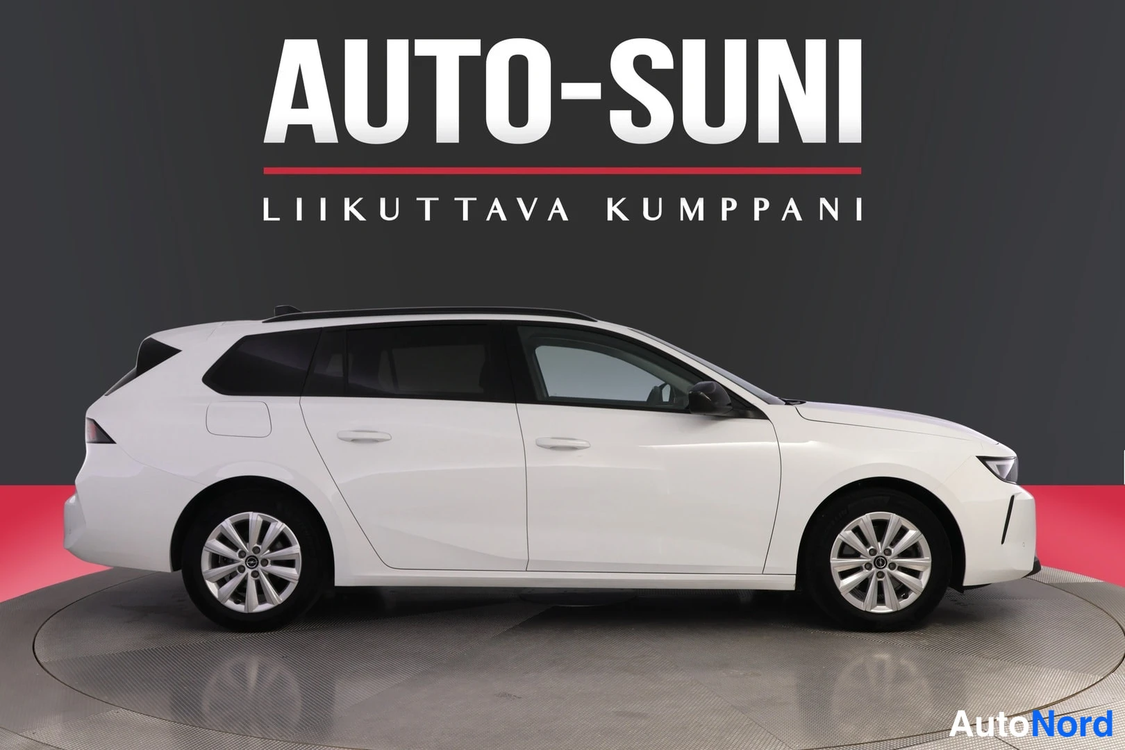 Sivulta: Valkoinen farmariauto sivulta kuvattuna studiossa. Näkyvät molemmat sivuosan ovet, tummat ikkunat, kattokaiteet ja hopeiset vannepyörät.