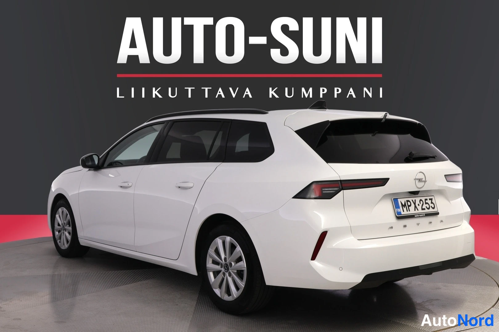 Takaviisto: Valkoinen farmariauto takakulmasta oikealta kuvattuna studiossa. Näkyvät takavalot, takaluukku Opel-tunnuksella ja Astra-tekstillä, rekisteri sekä auton vasen s
