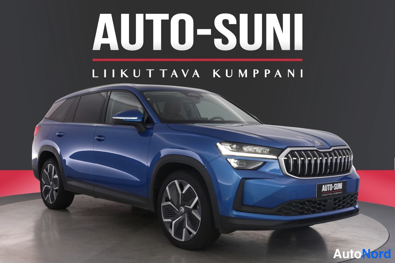 Etuviisto, nokka oikealle: Sininen tilavaauto studio-tyylisessä kuvassa, edessä oikealta kuvattuna, tausta tumma mainoskuvioinnilla Auto-Suni.
