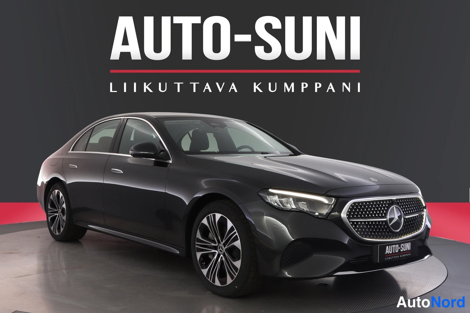 Etuviisto, nokka oikealle: Tummanharmaa Mercedes-Benz E-sarjan sedan näkyvissä etukulmasta oikealta, käännettynä näyttämöllä Auto-Sunin logotuin taustalla.