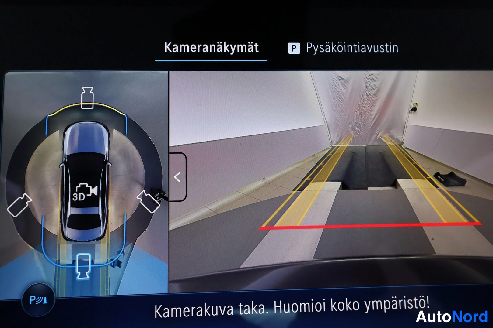 Edestä: Auton näytön kameranäkymä: 3D-kaava vasemmalla ja takakameran kuva pysäköintiviivoineen, tekstinä 'Kamerakuva taka. Huomioi koko ympäristö!'.