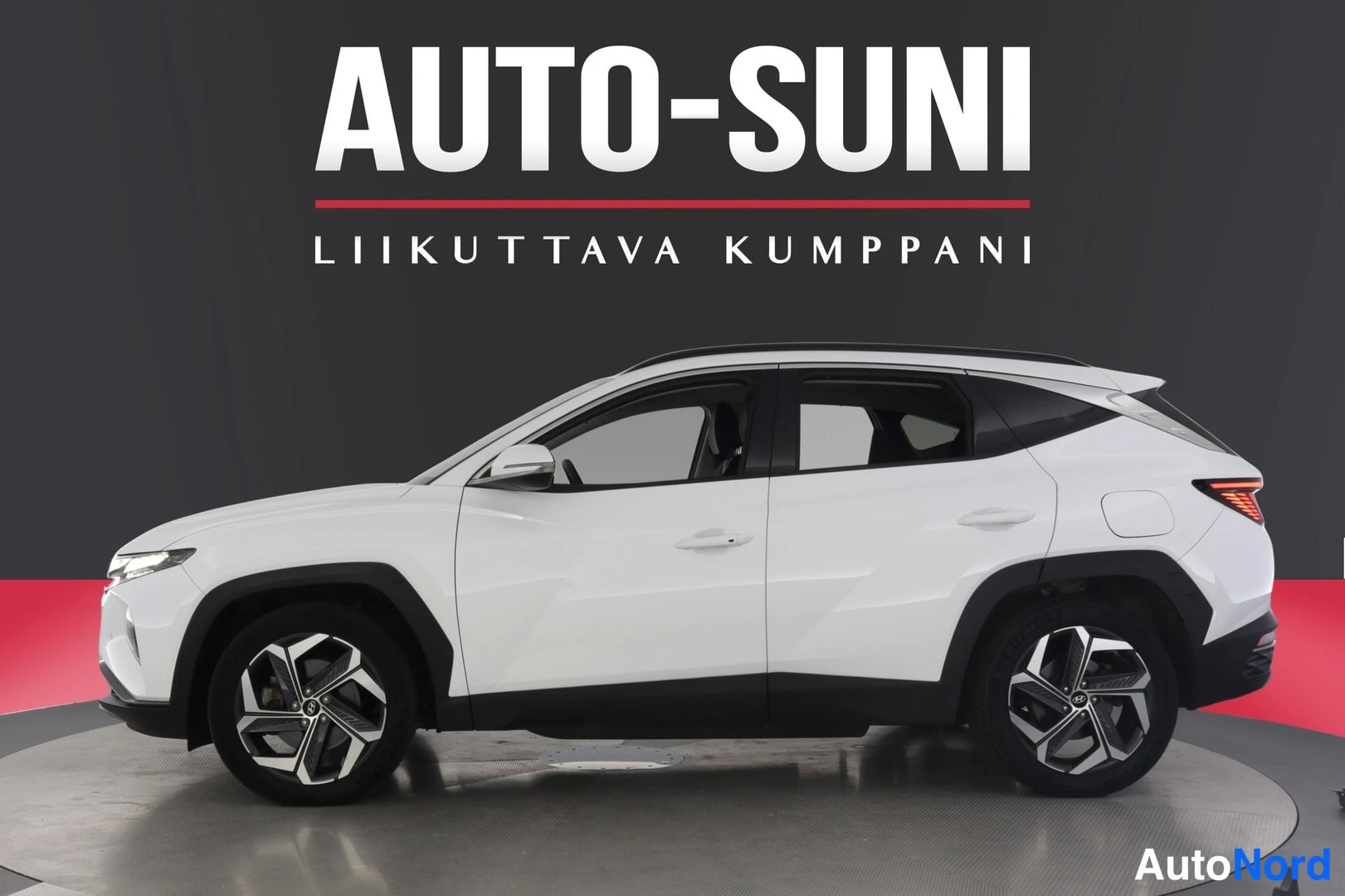 Sivulta: Valkoinen Hyundai Tucson nähty sivulta studio-ottelussa, mustat korokkeet ja AUTO-SUNI-taustakyltti.
