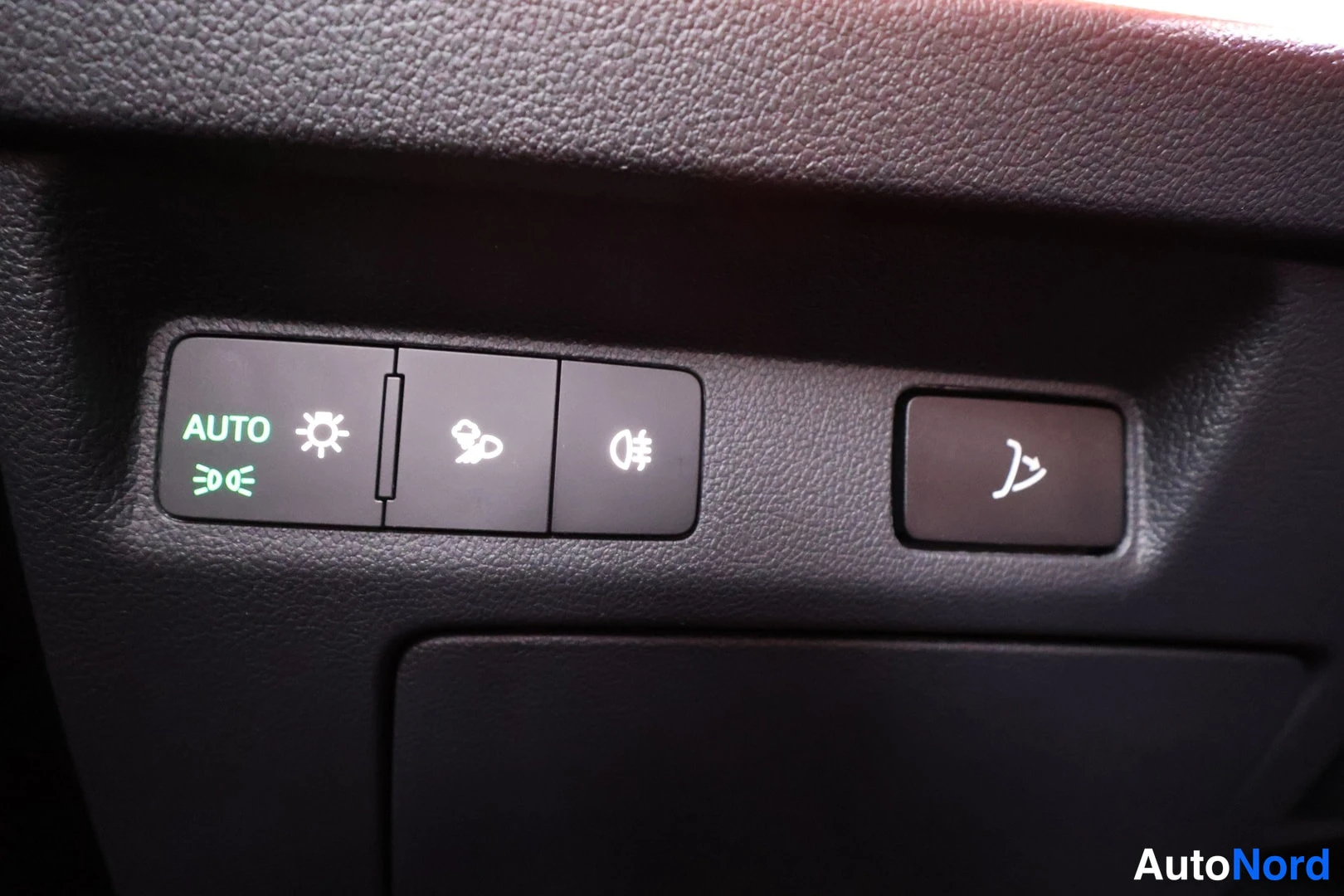 Edestä: Ajovalon kytkinpaneeli mustassa kojelaudassa, AUTO-teksti ja valokytkimet sekä tumma taustavaloa säätävä nappi. Kytkimissä lukuvat valaistut merkit suuntavaloje