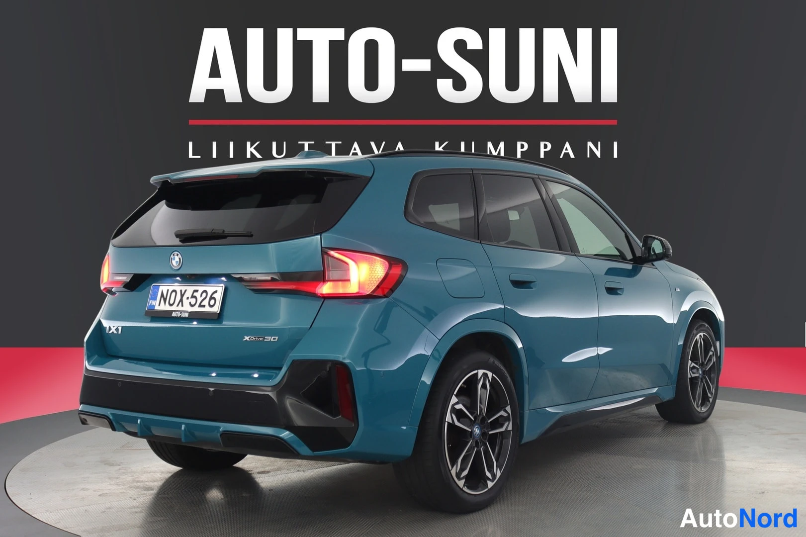 Takaviisto: Sinivihreä BMW iX1 -maasturi takaa studiotaustaa vasten, takavalot palavat.